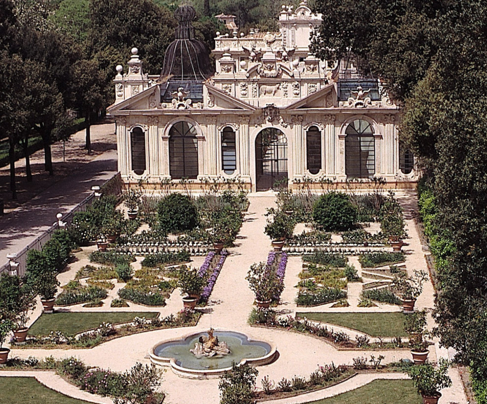 A symmetrical geometric garden with flower beds, paths, and a central fountain. A wrought-iron and stone fence separates the garden from a tree lined path at the left, and an ornate building with large windows and sculputral elements stands at the end of the garden.