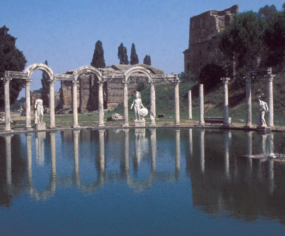 Roman ruins at Hadrian's Villa with stone columns and statues reflected in a pond, with trees and old masonry buildings in the background under a clear blue sky.