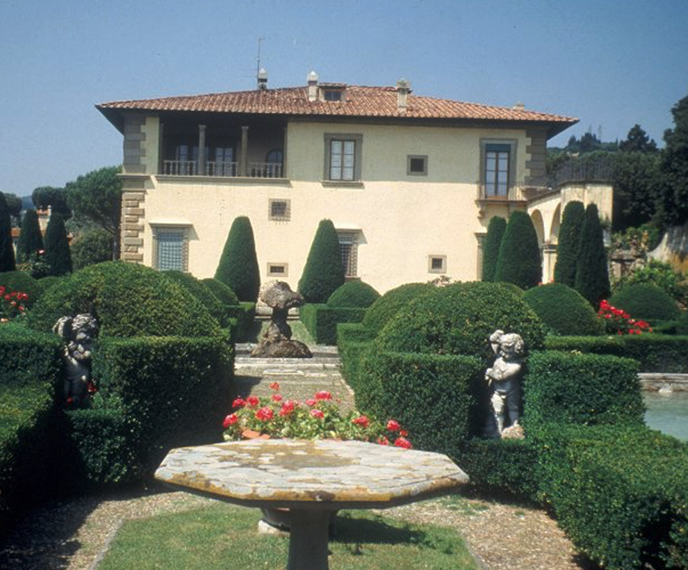 A neatly manicured garden with sculpted hedges, rose bushes, and cherub statues, in front of a two-story villa with a red tile roof under a clear blue sky.
