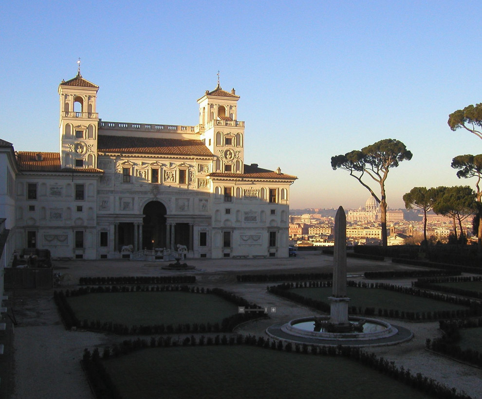 A grand symmetrical building with two towers stands behind a formal garden with geometric hedges and a central obelisk, overlooking a cityscape.