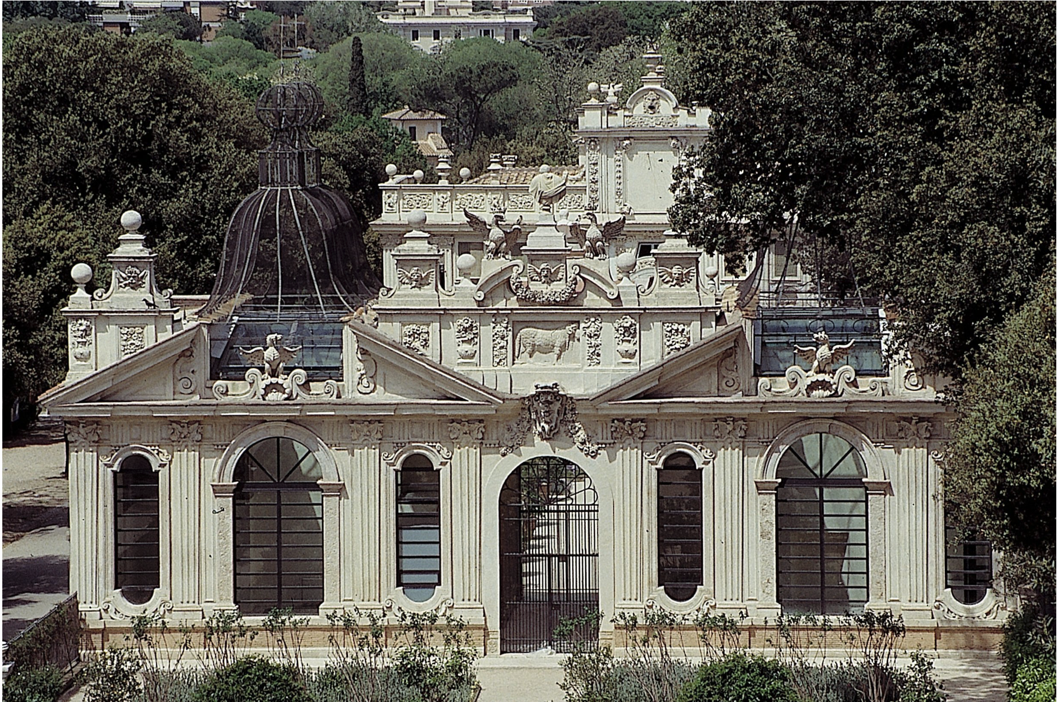 A large stone structure with arched windows and carved decorative sculptures. A wrought-iron gate fills the arched entryway, and trees and vegetation appear in the foreground and background.