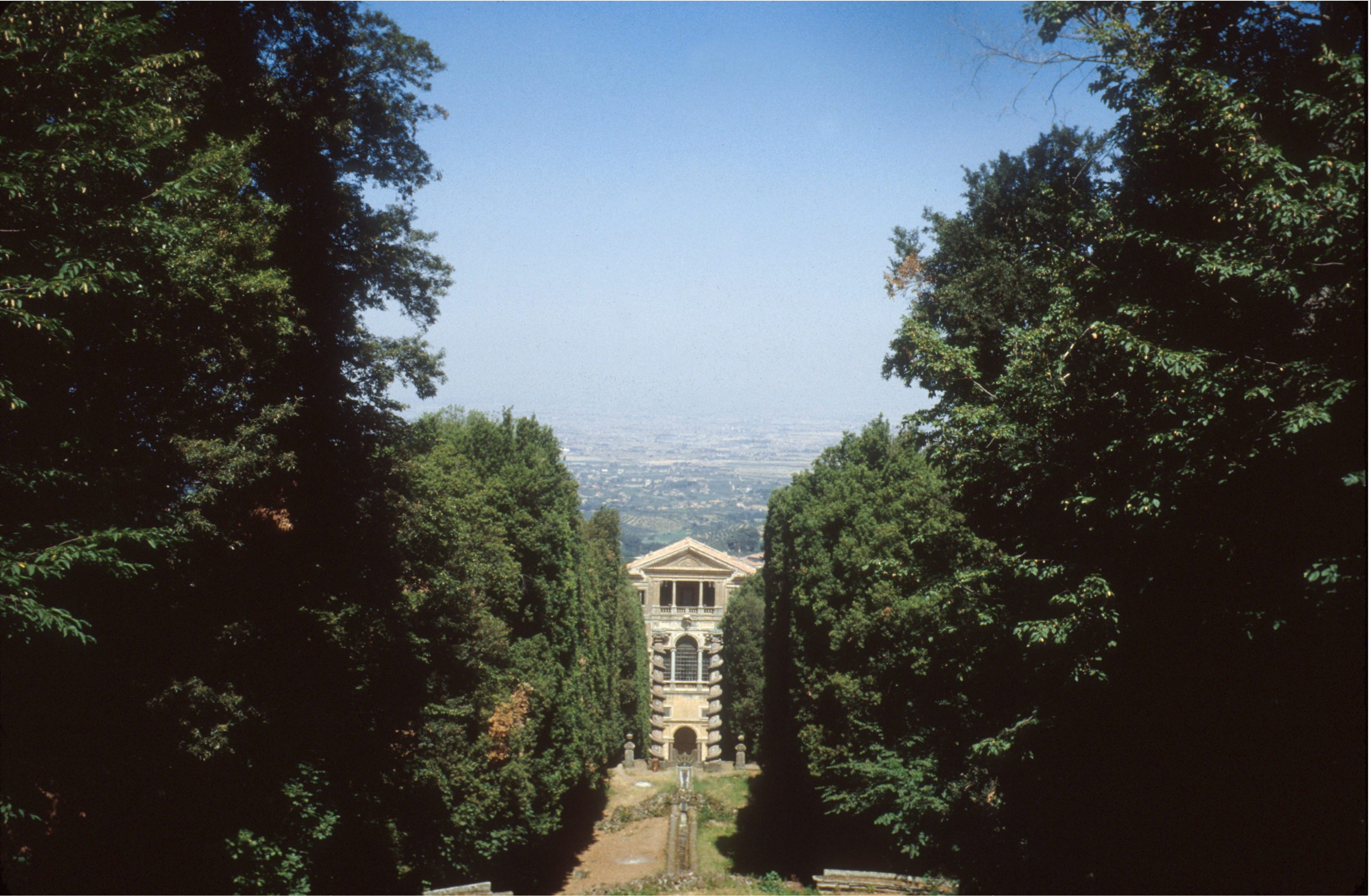 A stone building stands at the end of a tree-lined path, framed by tall, dense greenery on both sides, with a hazy cityscape visible in the distance under a clear blue sky.