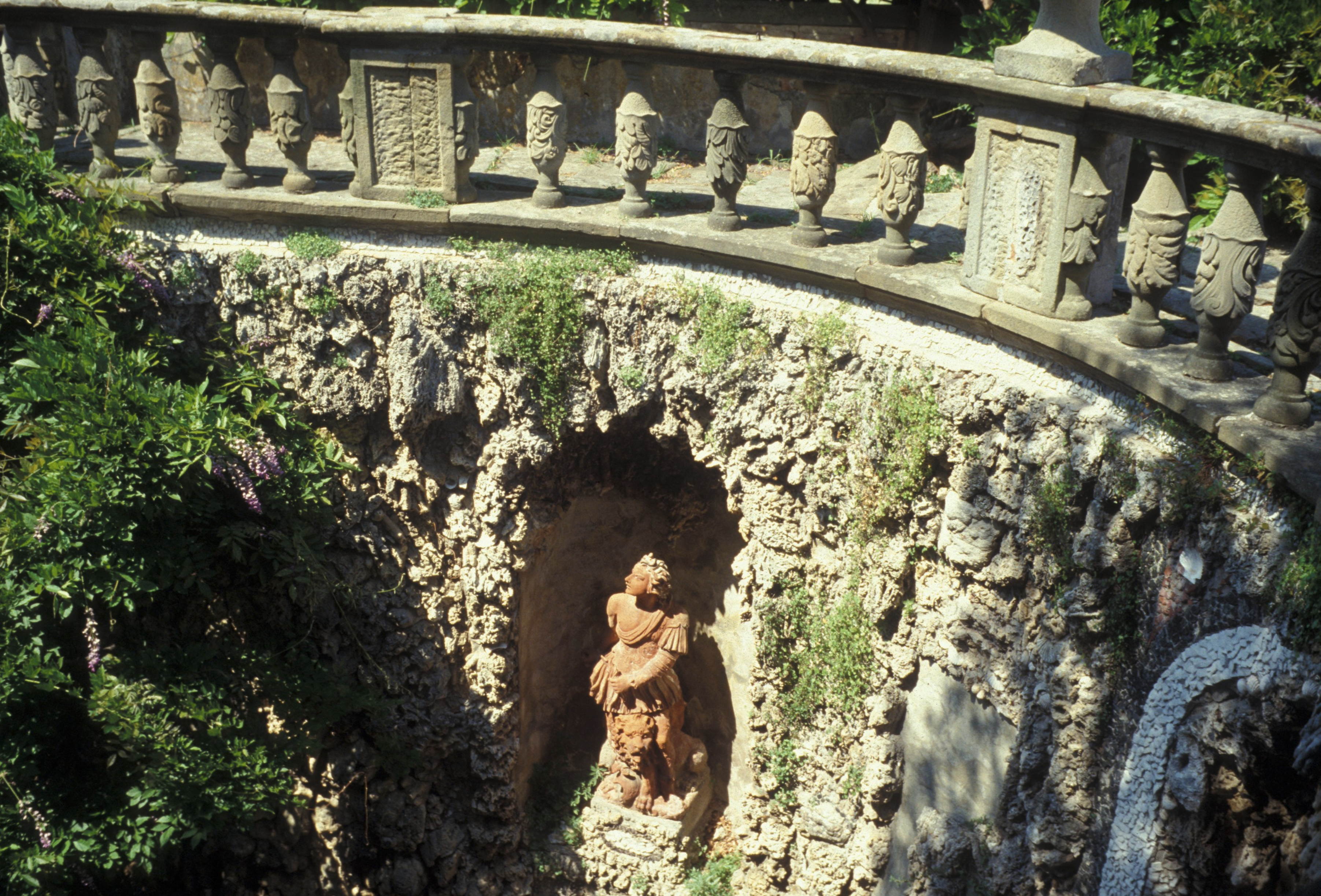 A view from above of rocaille decorated wall covered in moss and stone, with a terracotta-colored statue in an alcove. A balustrade lines the top of the wall and greenery appears in the foreground and behind the balustrade.