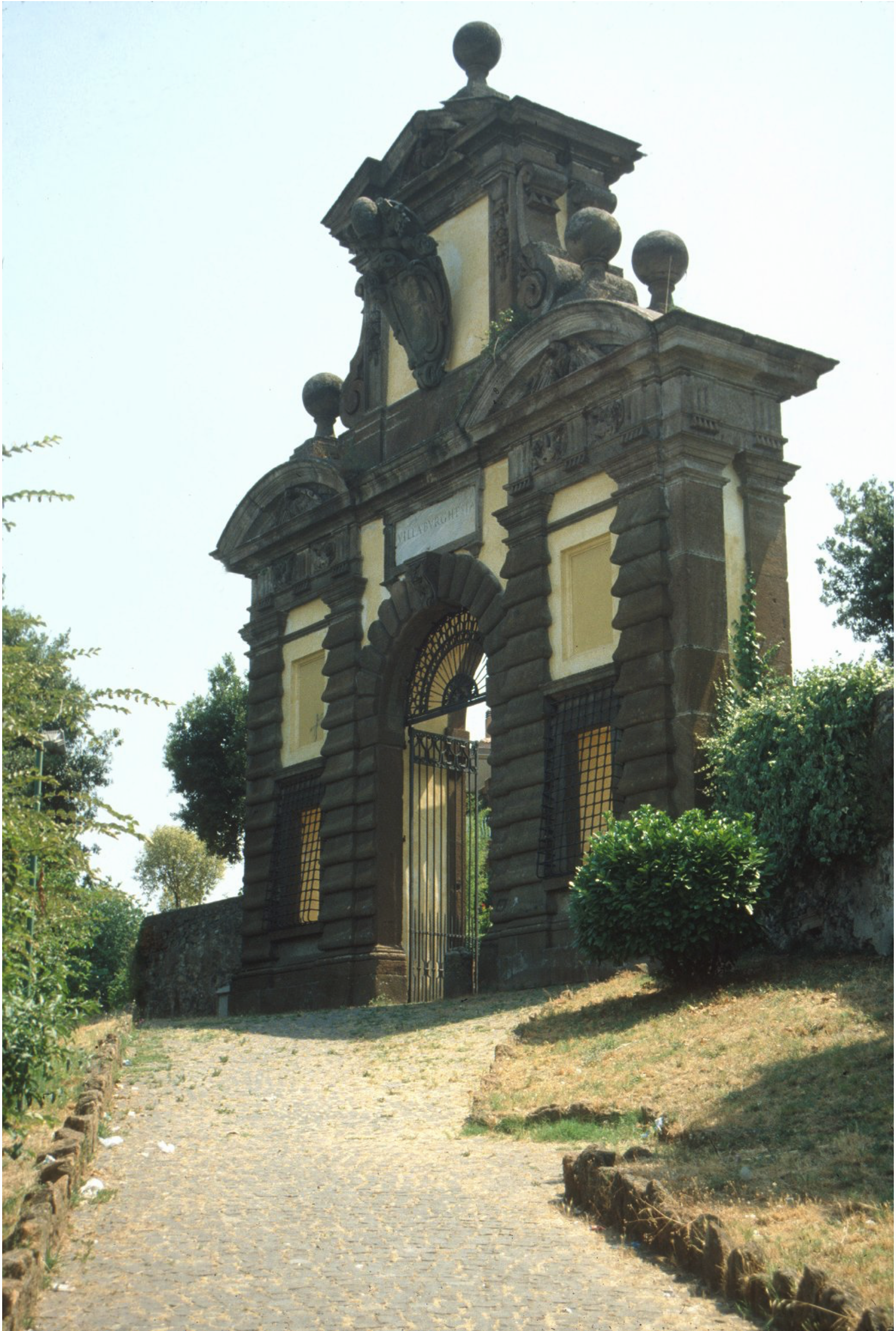 A stone archway with a central gate, topped with decorative sculptural elements, stands at the end of a cobblestone path, surrounded by greenery and trees.