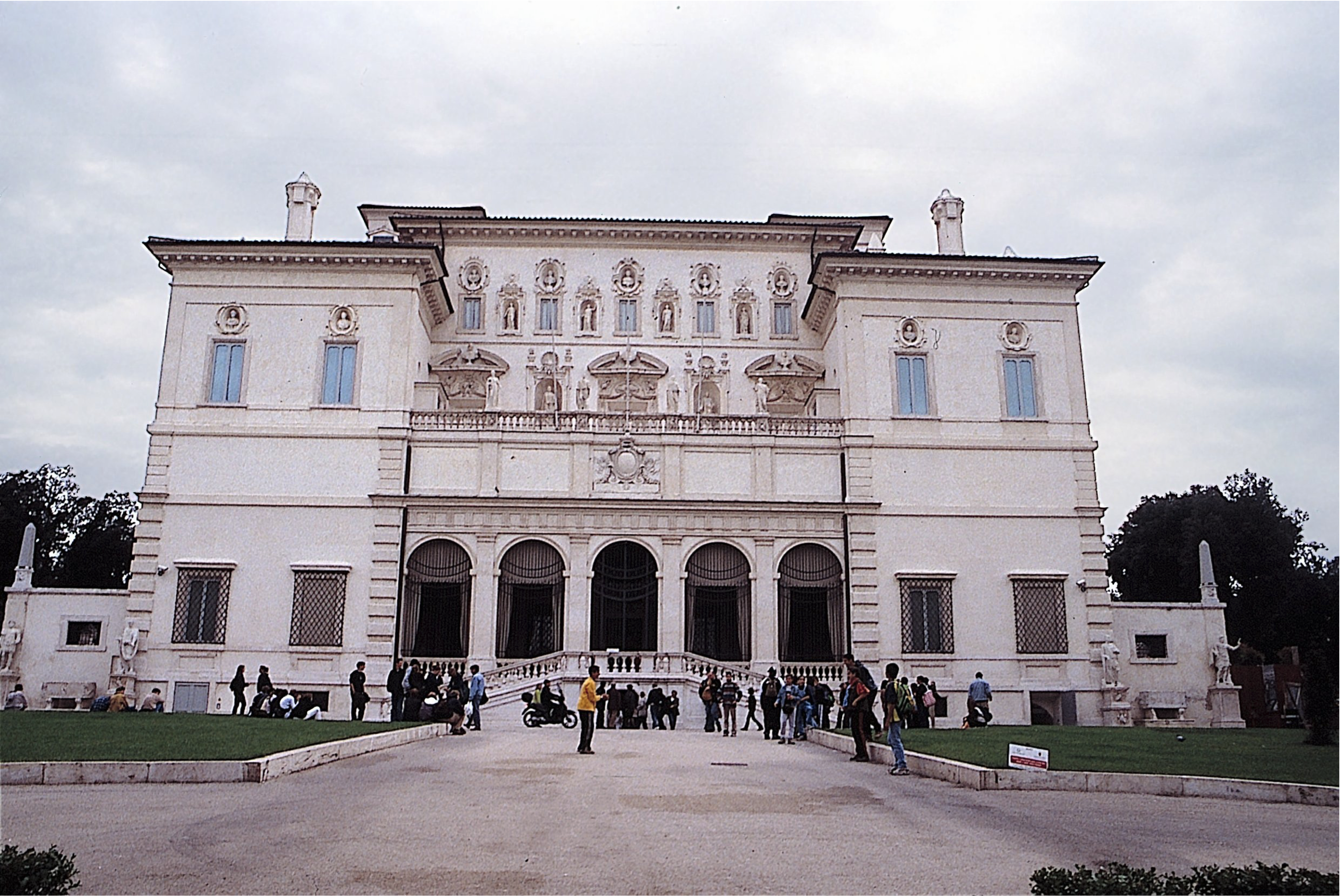 A large, ornate villa with detailed carvings and statues on its symmetrical facade. People are gathered outside the entrance, and there is a wide path with grass on either side leading up to the building.