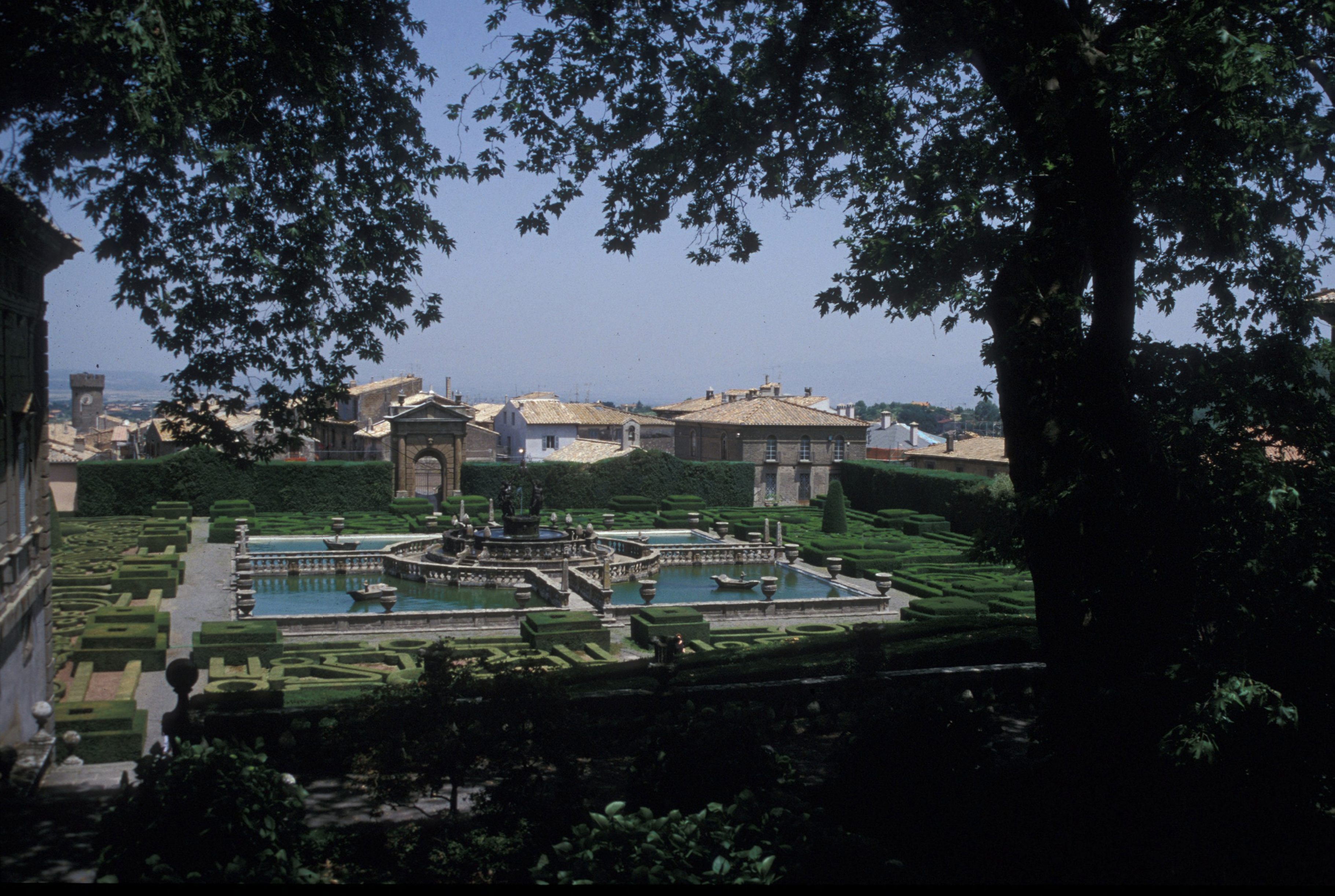 A formal garden with ornate fountains, geometric hedges, paths and reflecting pools. Stone buildings and tiled rooftops are visible in the background, while tree branches frame the foreground.