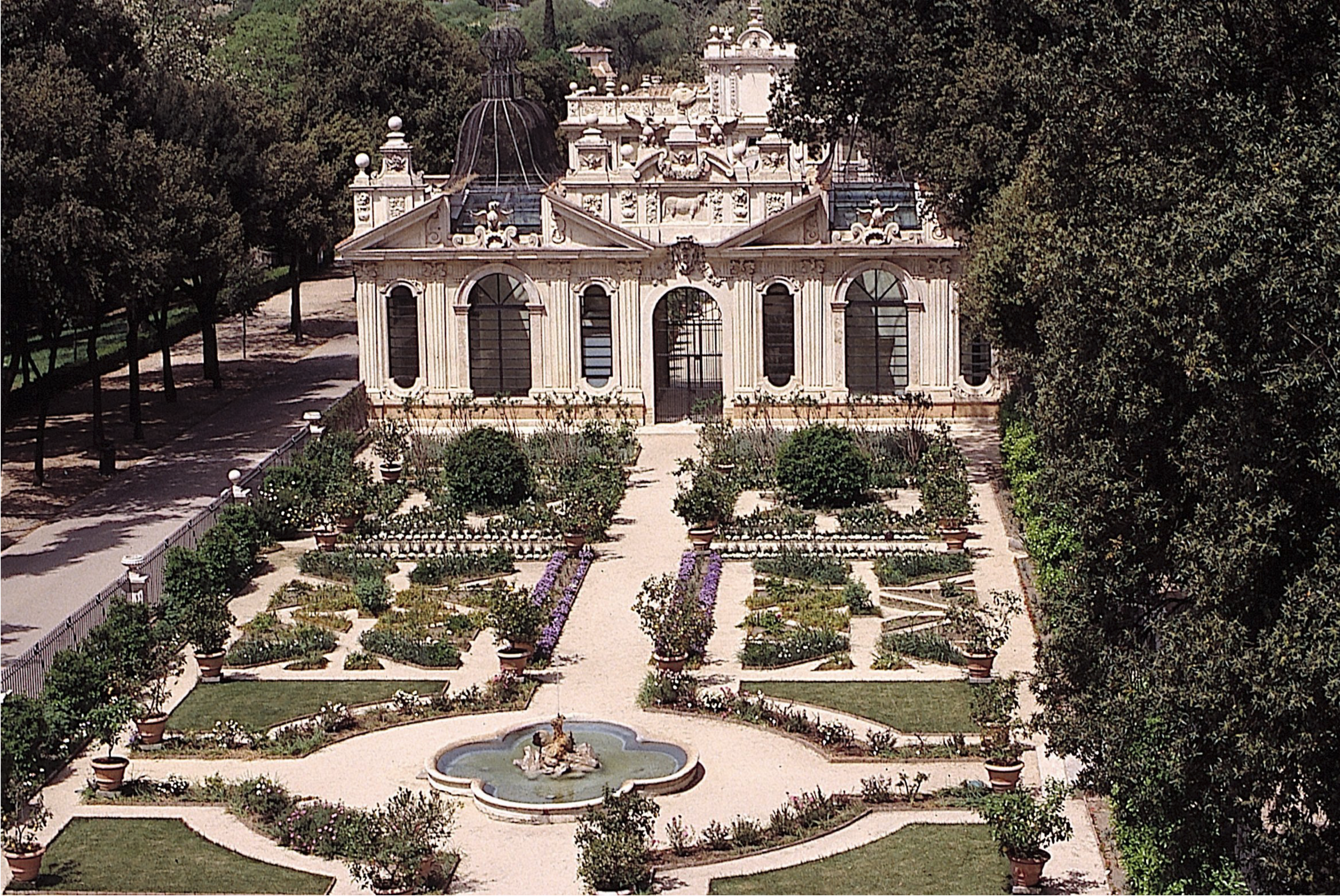 A symmetrical geometric garden with flower beds, paths, and a central fountain. A wrought-iron and stone fence separates the garden from a tree lined path at the left, and an ornate building with large windows and sculputral elements stands at the end of the garden.