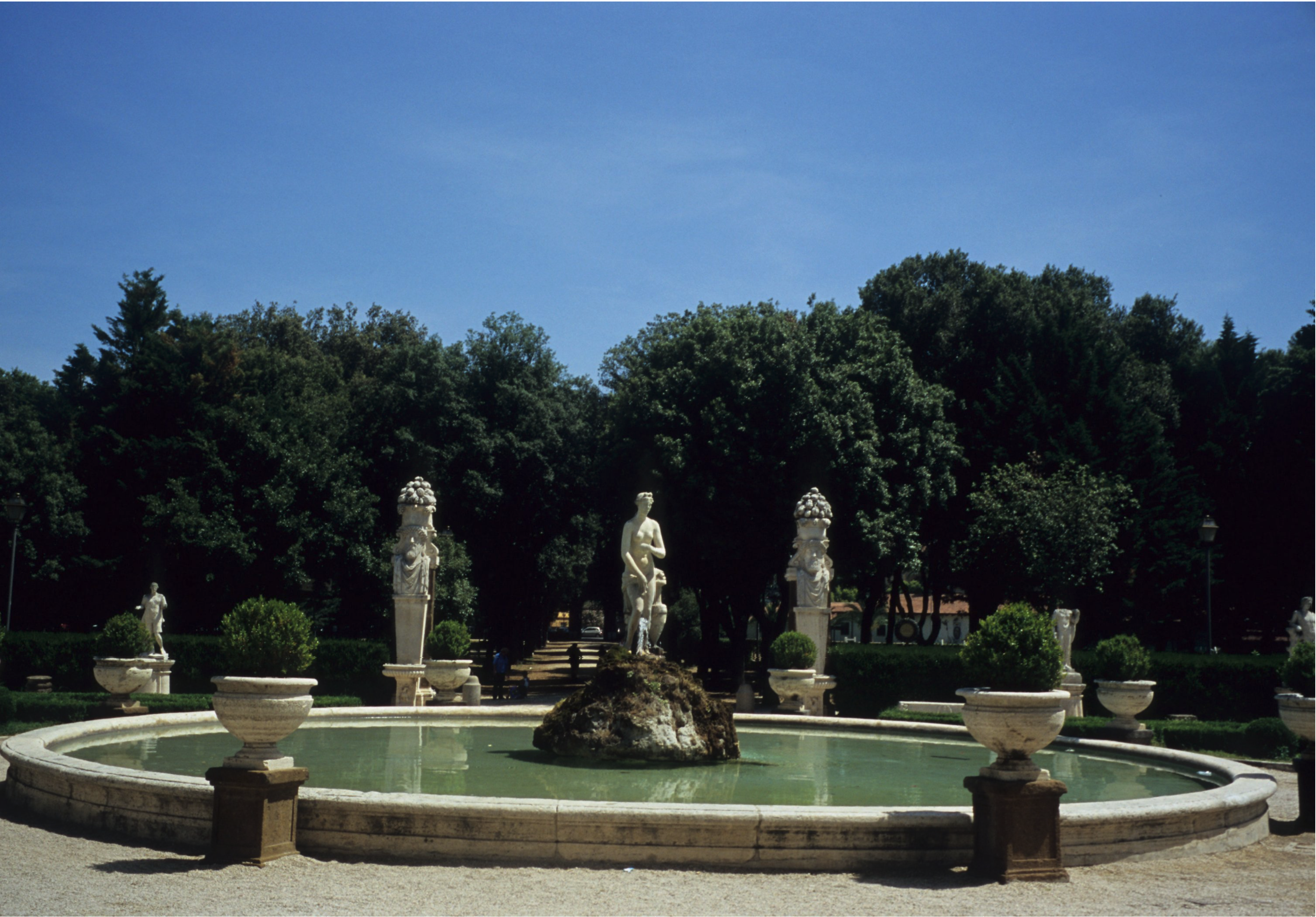A round stone fountain with a classical female statue on top of a rocky outcropping in the center. The fountain is surrounded by statues, urns, and hedges, with a shaded allée of trees in the background.