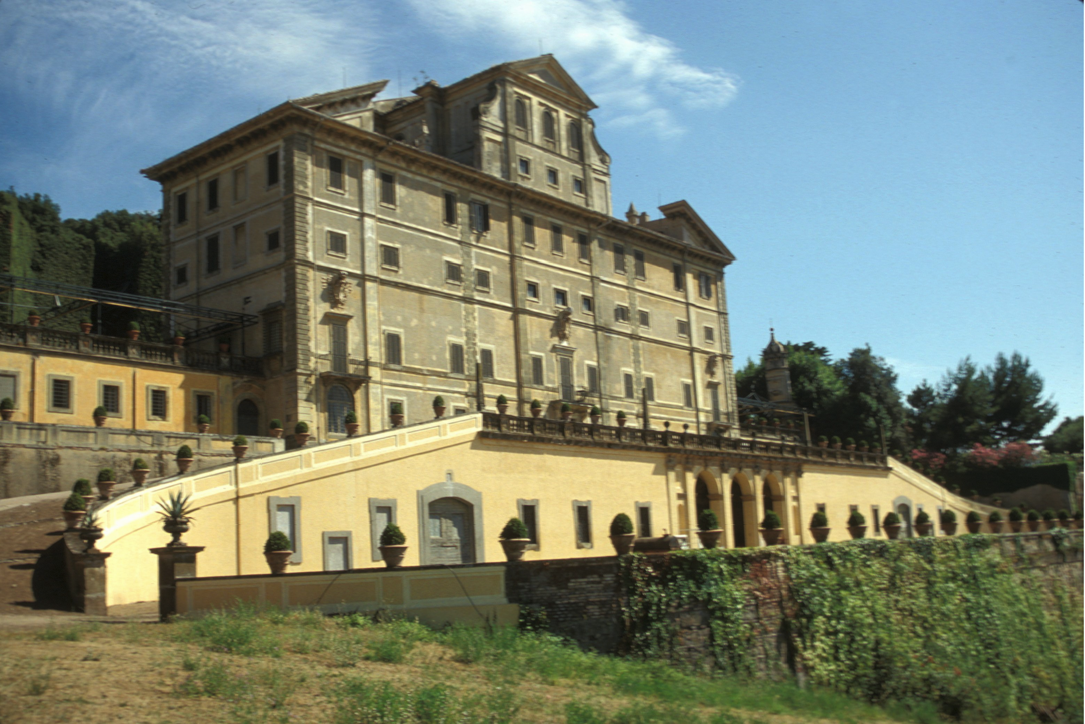 A large, historic villa with beige stone walls and rows of windows sits on a hill, with a terraced staircase, balustrades, potted plants, and green ivy growing along the lower wall under a blue sky.