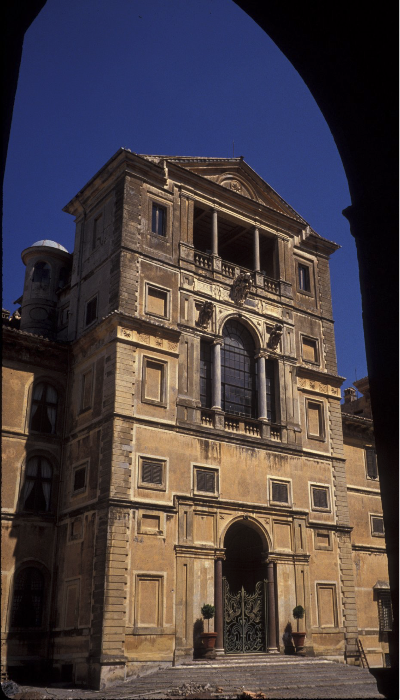 A multi-story stone building with decorative columns archways, and symmetric rectangular windows stands under a clear blue sky, viewed from a shaded archway in the foreground.