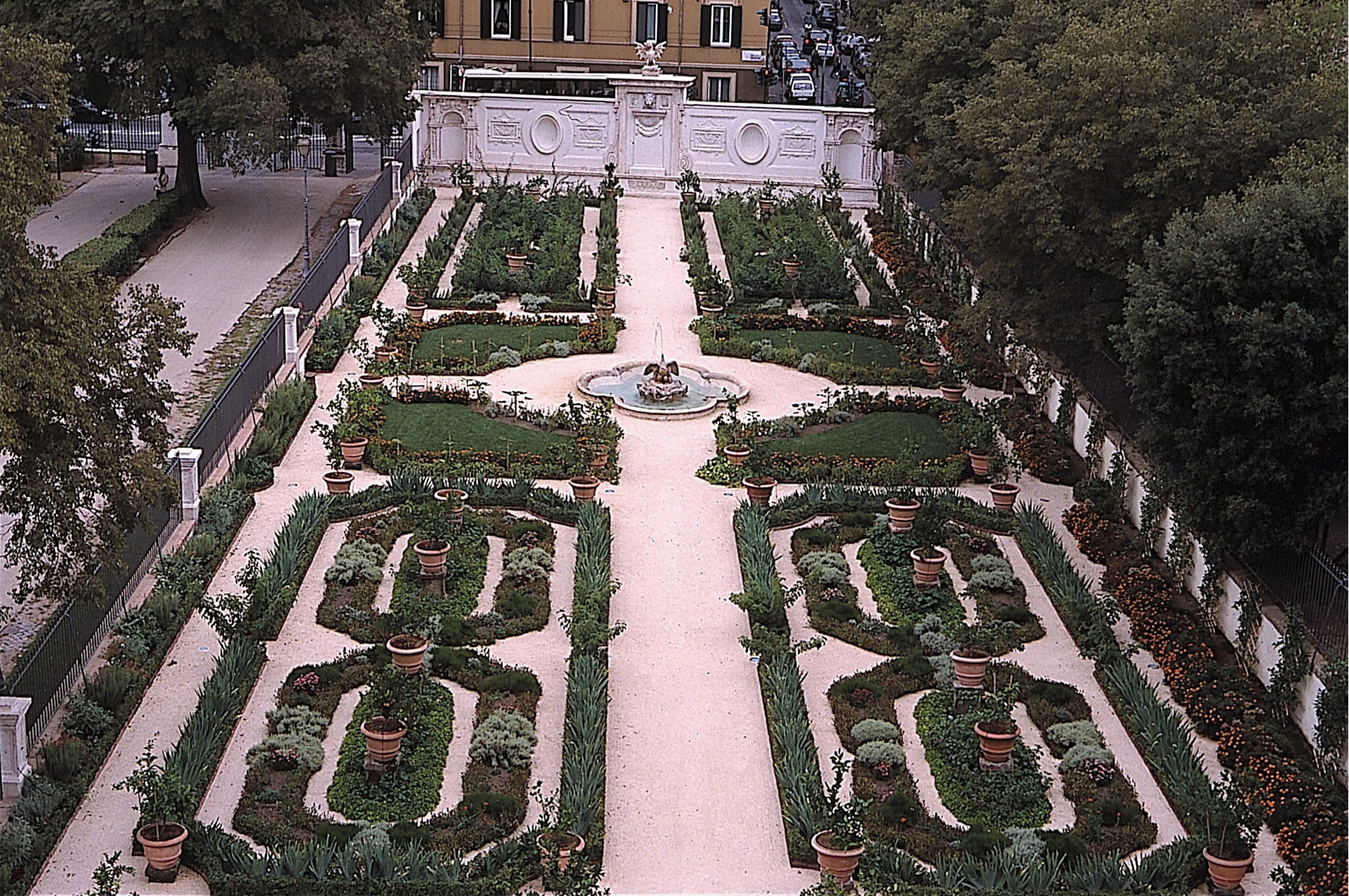 A formal garden with symmetrical geometric flower beds and paths and a central fountain; The garden is surrounded by an iron fence and ornamented wall, with urban streets and buildings in the background.