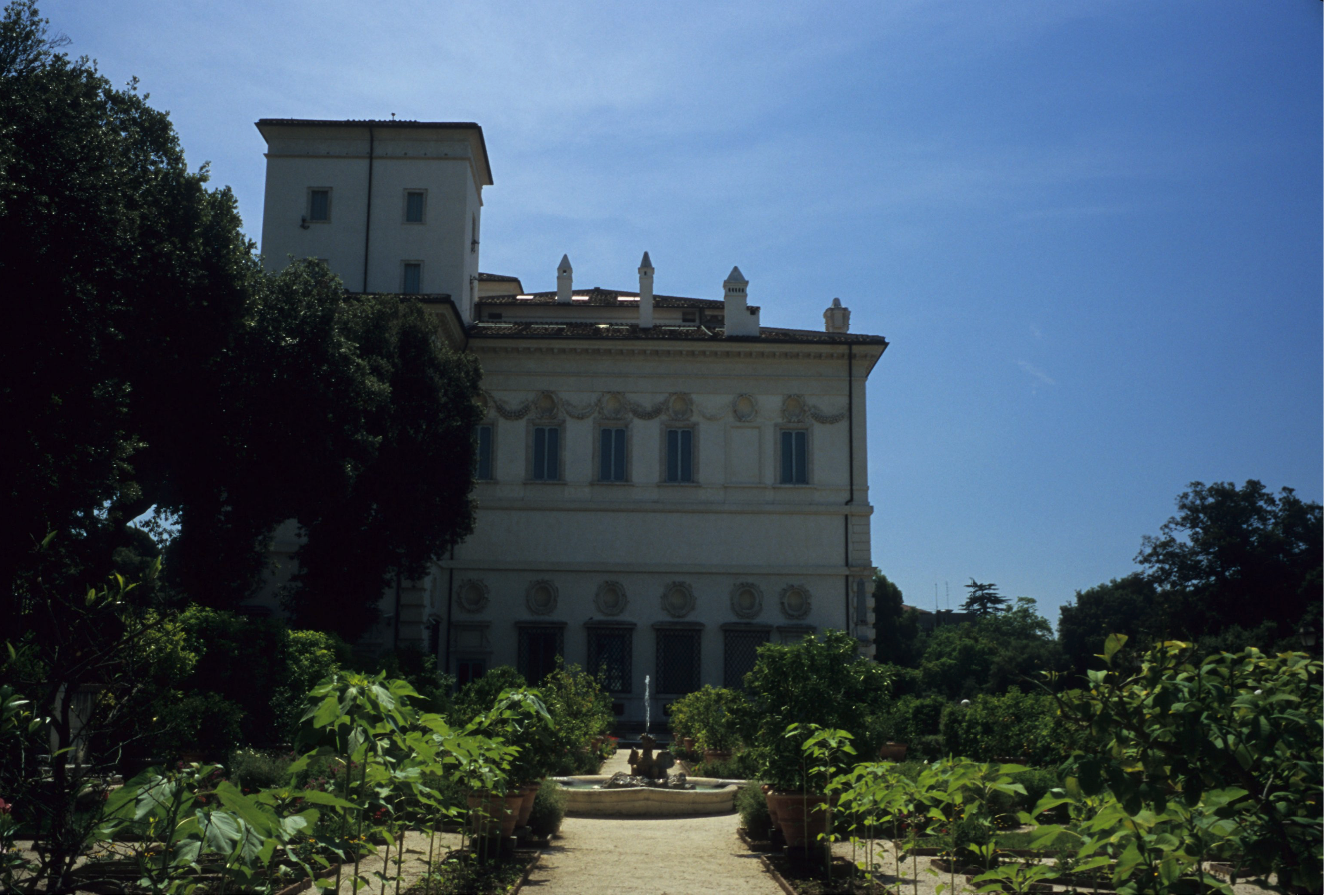 A grand villa with ornate architectural details stands at the end of a lush garden with paths, greenery, and fountain and under a clear blue sky.