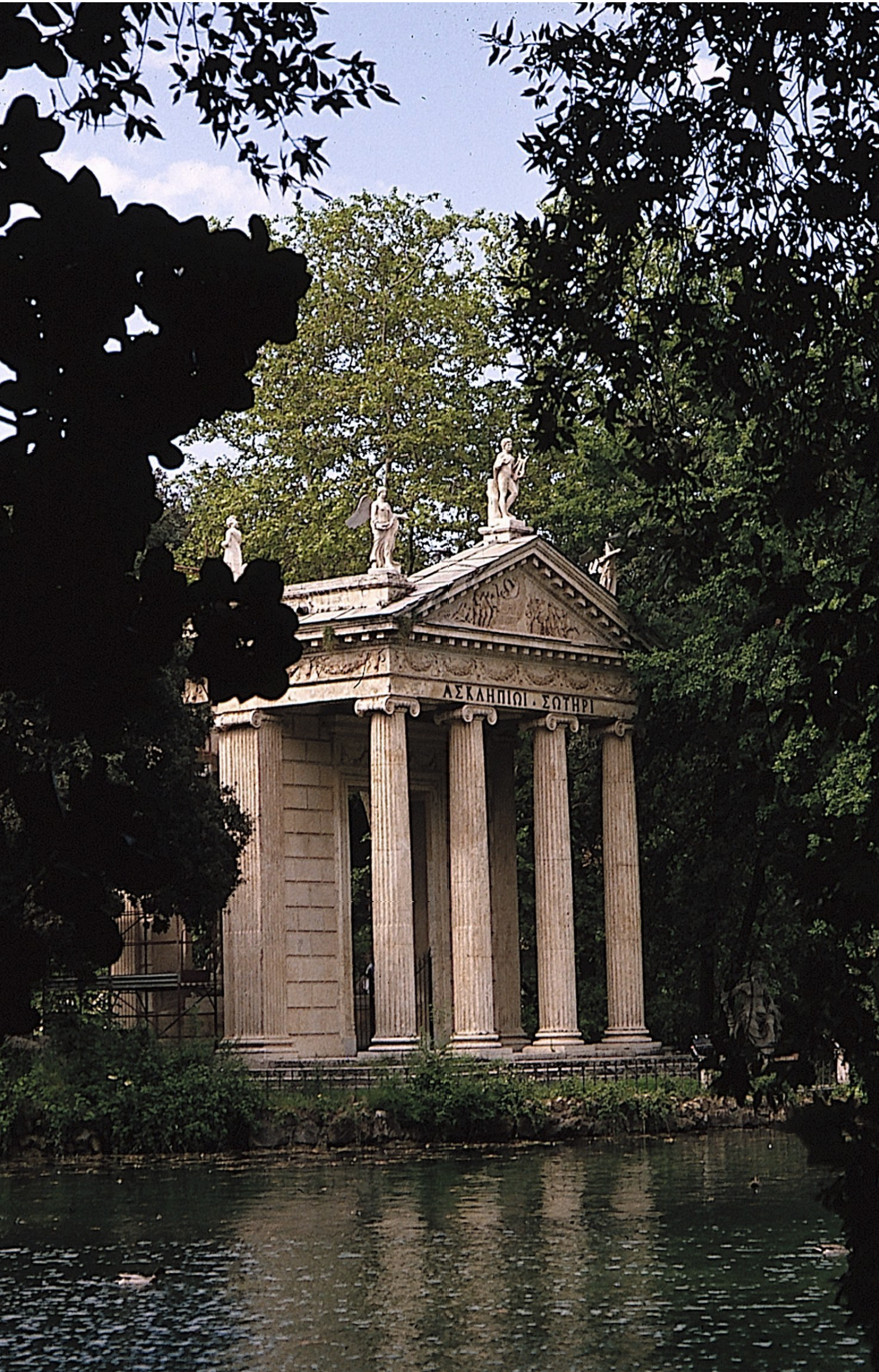 A classical-style temple with tall columns and a roof decorated with statues stands by a body of water, surrounded by lush green trees and foliage.