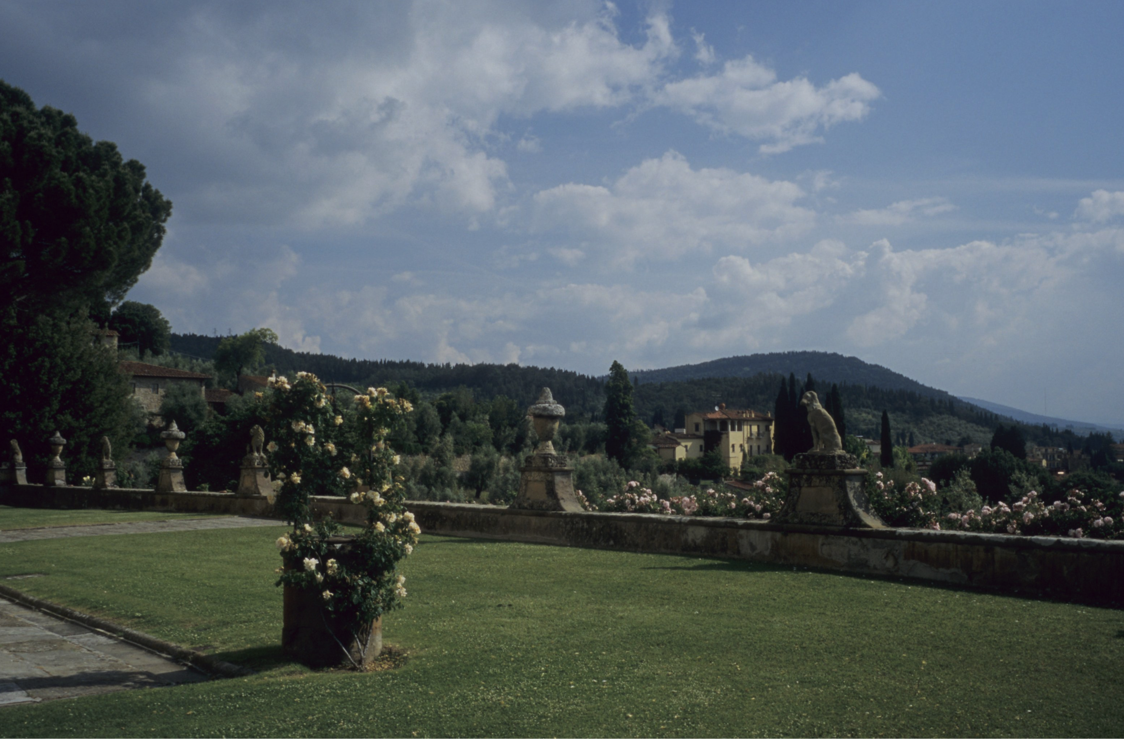 A scenic view of a lush, manicured lawn with stone statues, flowers and a low wall, overlooking rolling hills, trees, and distant buildings under a partly cloudy sky.