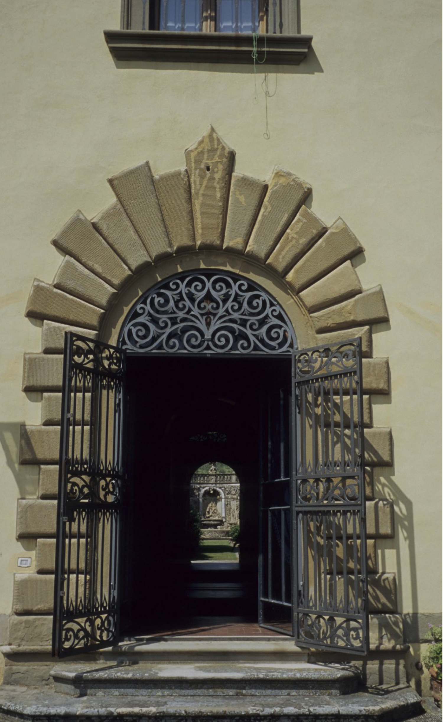 An arched doorway with decorative iron gates, set in a light stone building at the top of a set of stone steps, opens to reveal a corridor leading to a garden with greenery and a stone structure in the distance.