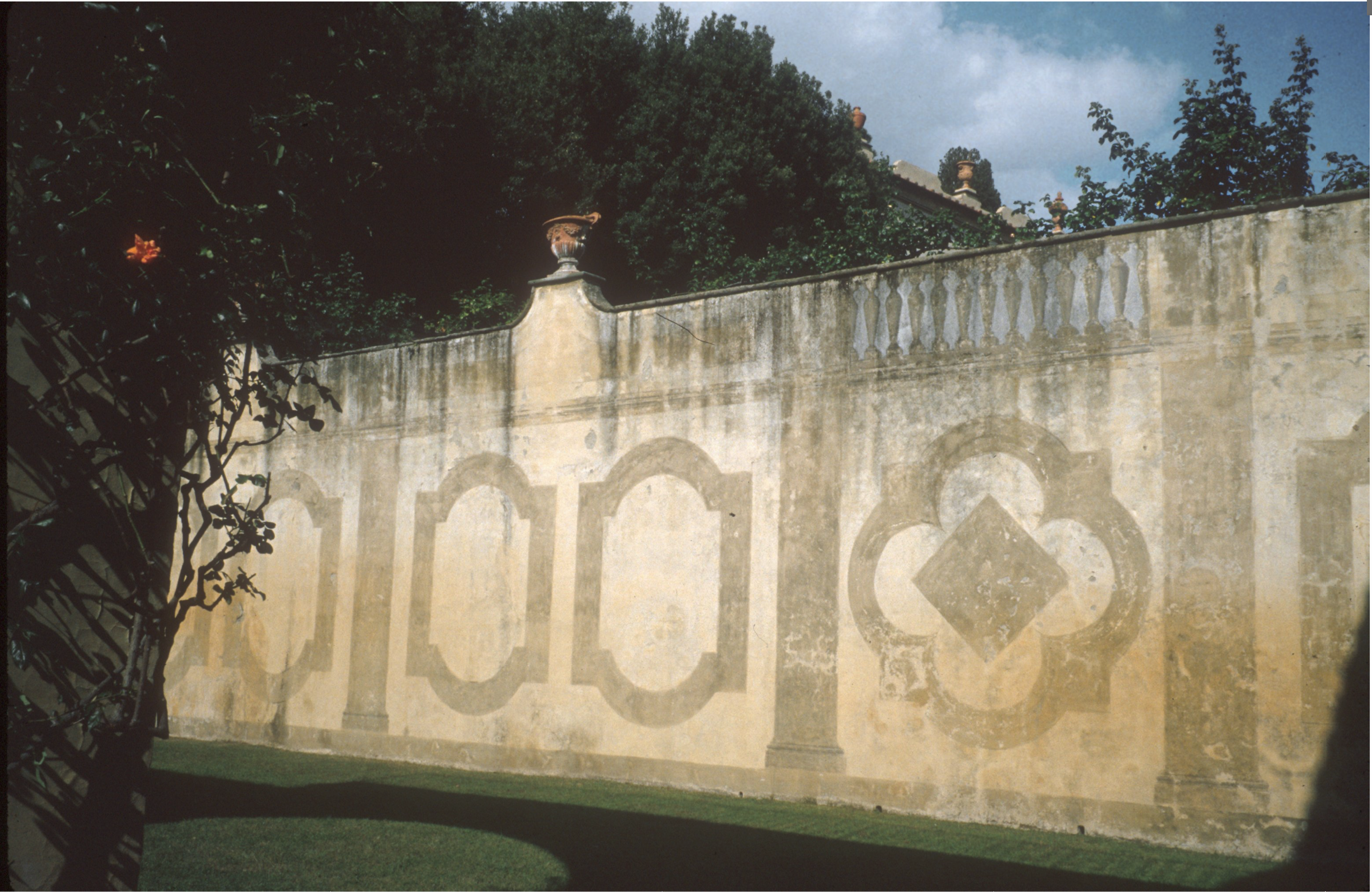 A garden wall with beige and cream geometric designs, decorative urns on top, and green trees visible behind the balustrade. Grass and some leafy branches are in the foreground.