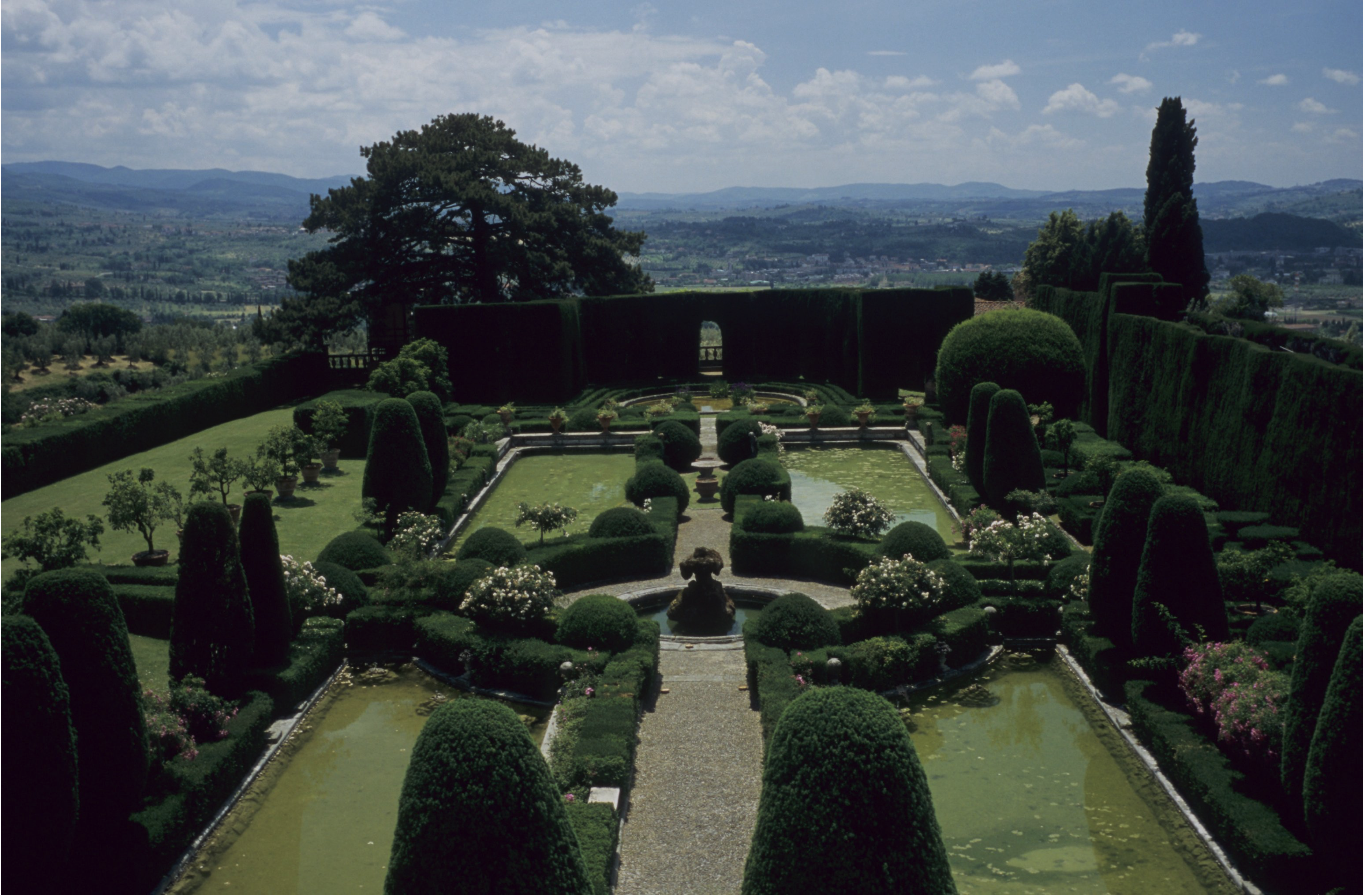 A formal garden with rounded hedges, symmetrical pathways, rectangular ponds and a central fountain, set against a backdrop of hills and a cloudy blue sky. Tall trees and a trimmed hedge wall border the garden.