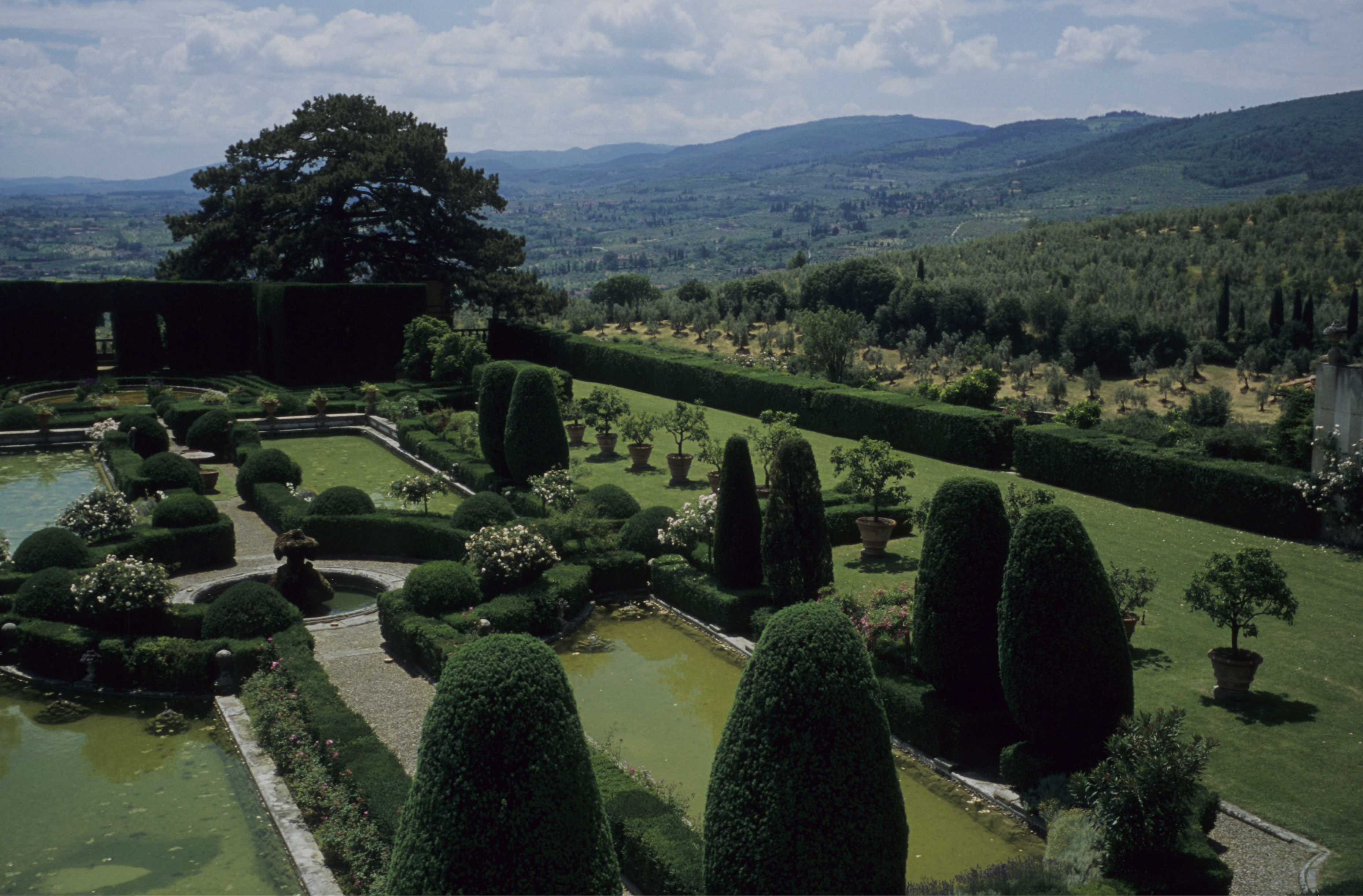 Aerial view of a formal Italian garden featuring rounded hedges, reflecting pools, and potted citrus trees, with a landscape of rolling hills and plains in the background.