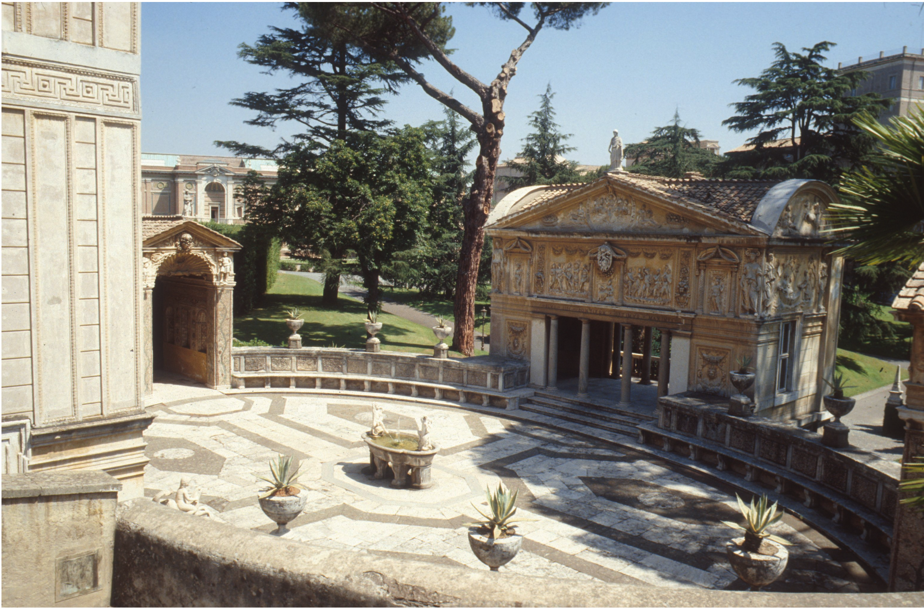 A courtyard with ornate stone buildings, patterned stone flooring, a central fountain, potted plants, and large trees in the background.