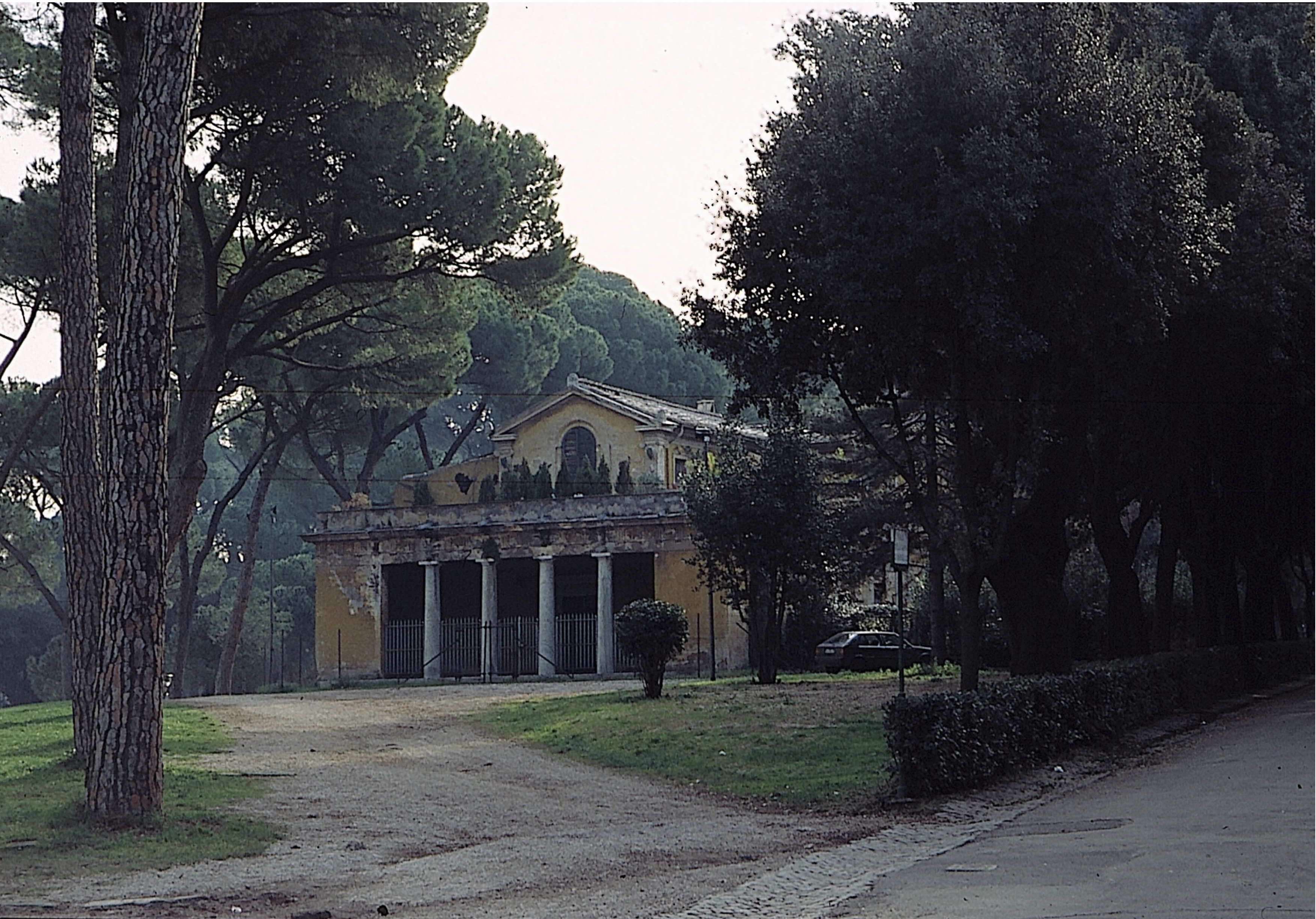 A yellow building with a porch and stone columns behind an iron fence stands among tall trees and greenery along a dirt path, with a parked car partially visible beside it.