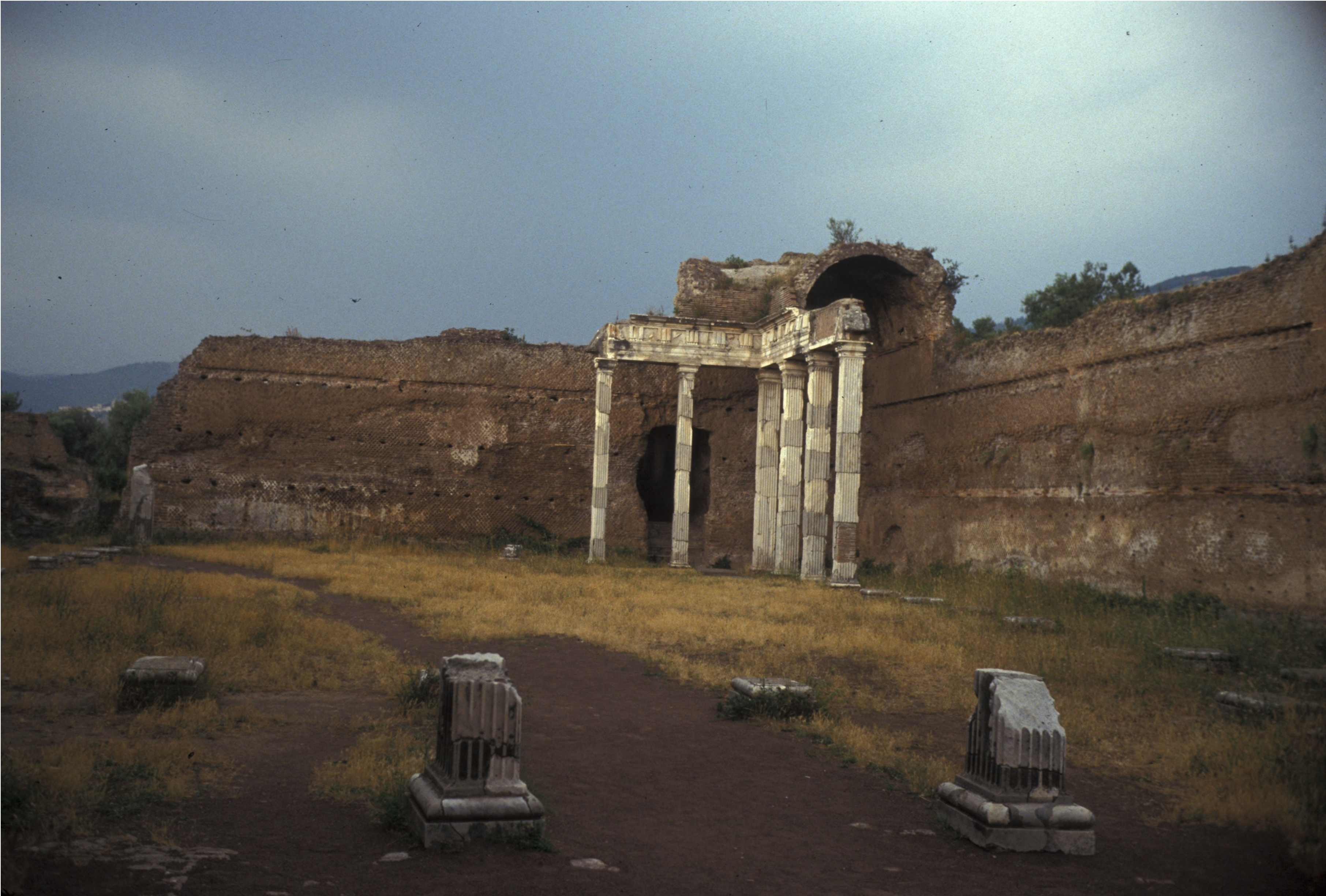 Stone ruins of a partially standing archway and columns, surrounded by tall, weathered brick walls and scattered stone fragments on a grassy, dirt-covered ground.