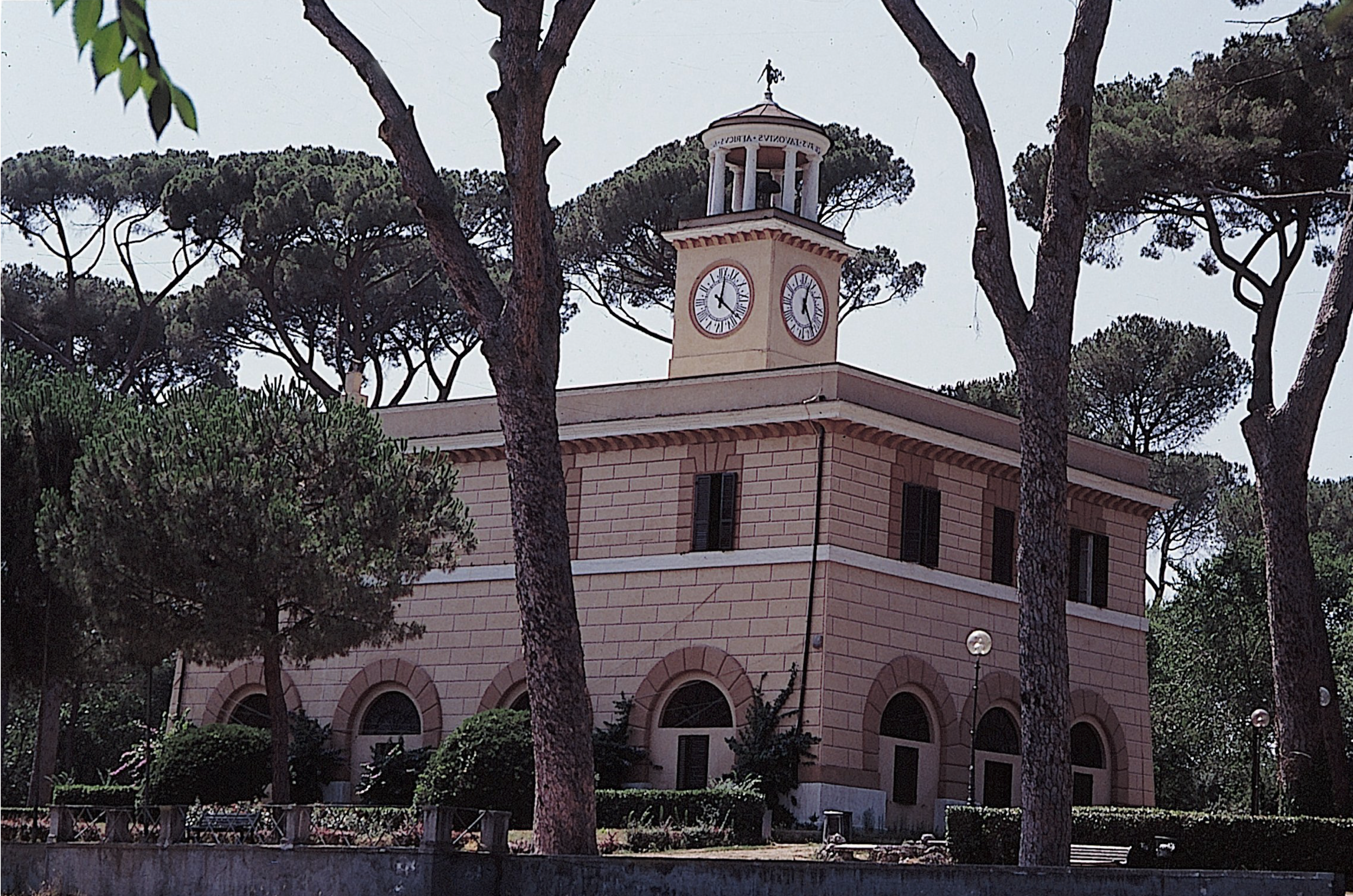 A two-story, beige-colored building with rectangular and arched windows and a clock tower sits among tall pine trees, surrounded by greenery.