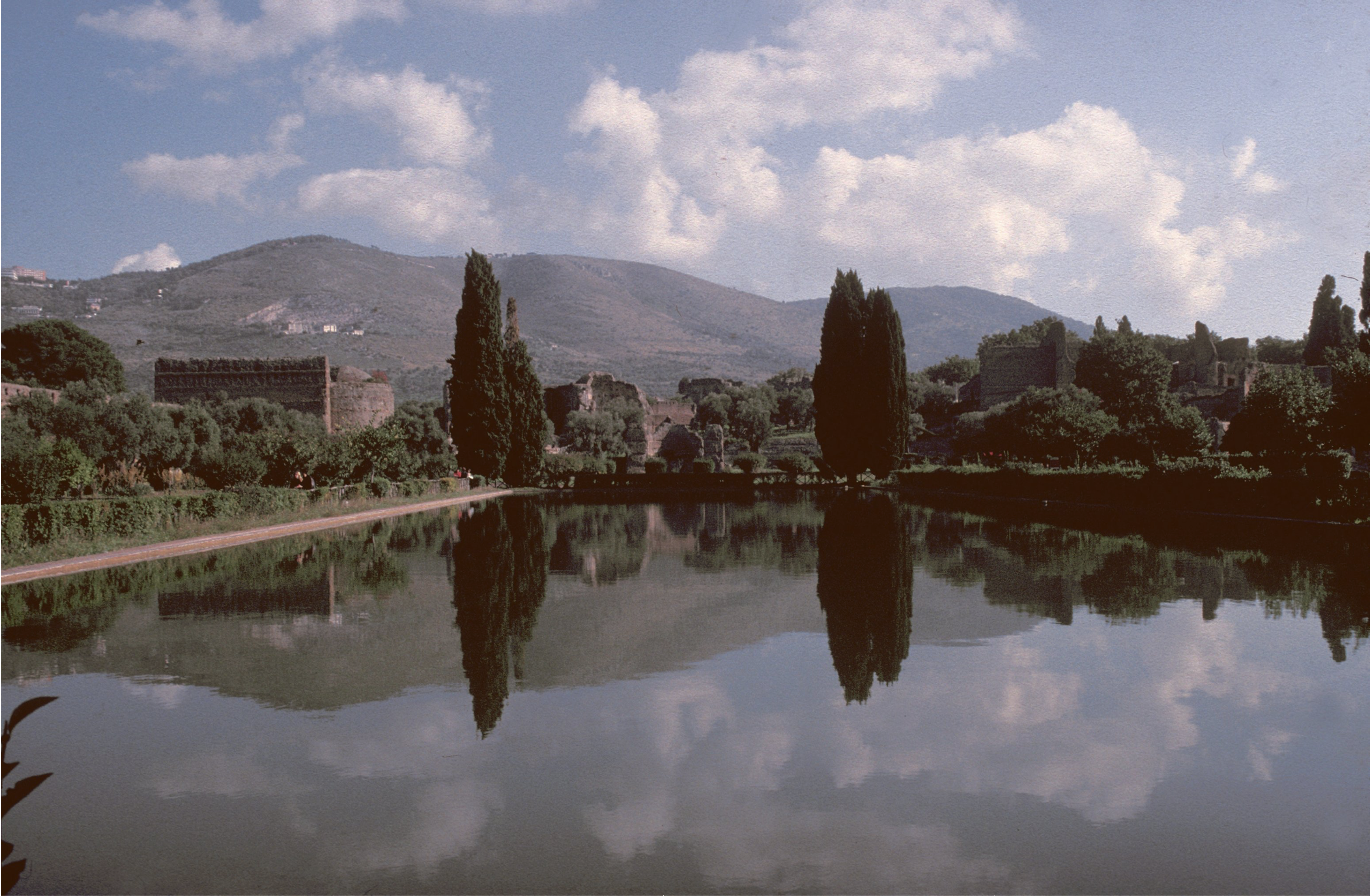 A large rectangular reflecting pool surrounded by trees and greenery, with distant hills and a partly cloudy sky mirrored in the water. Brick structures are partially visible among the trees.
