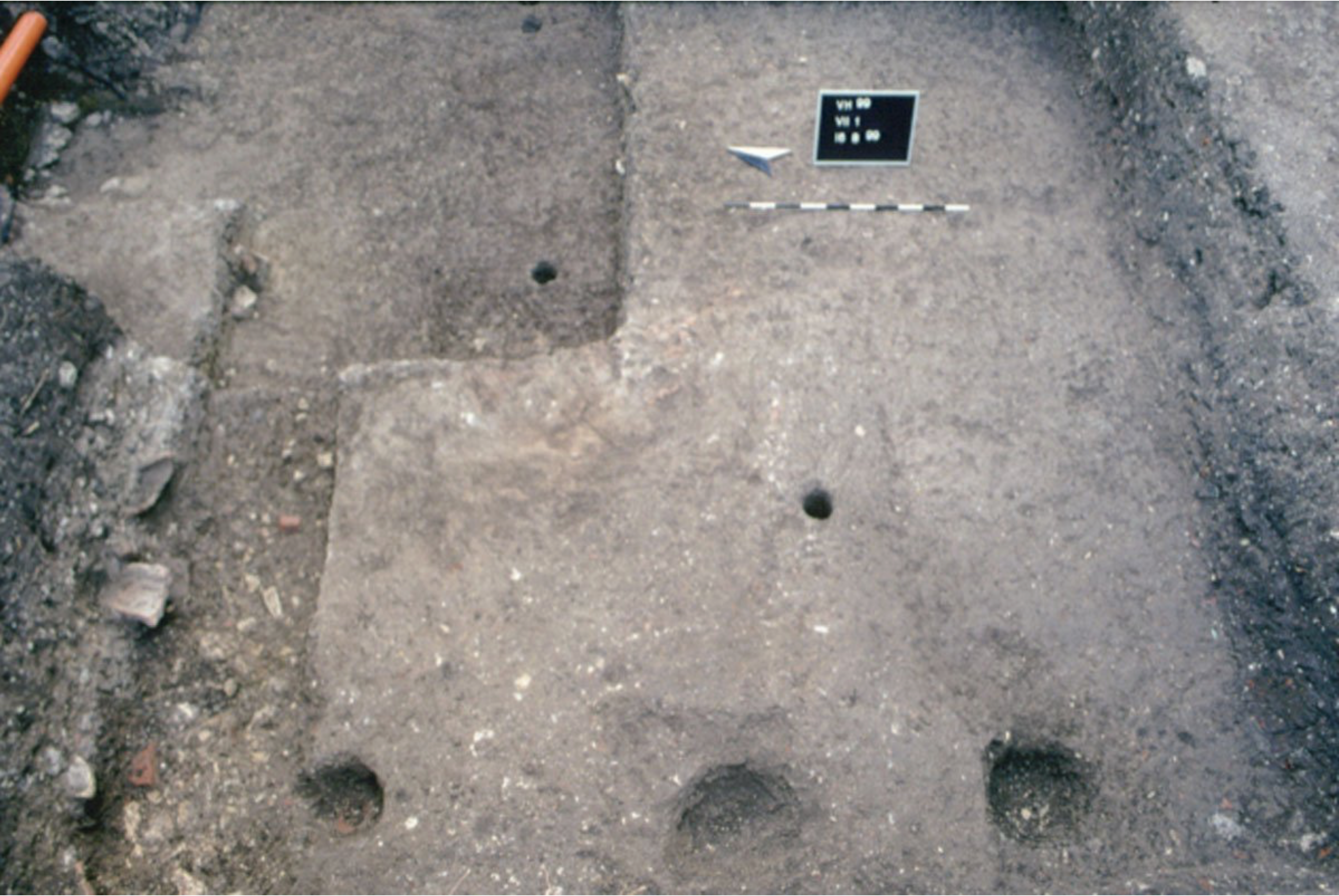 A rectangular archaeological excavation site with several circular post holes in the ground and a measuring scale and information board placed near the top center of the image.