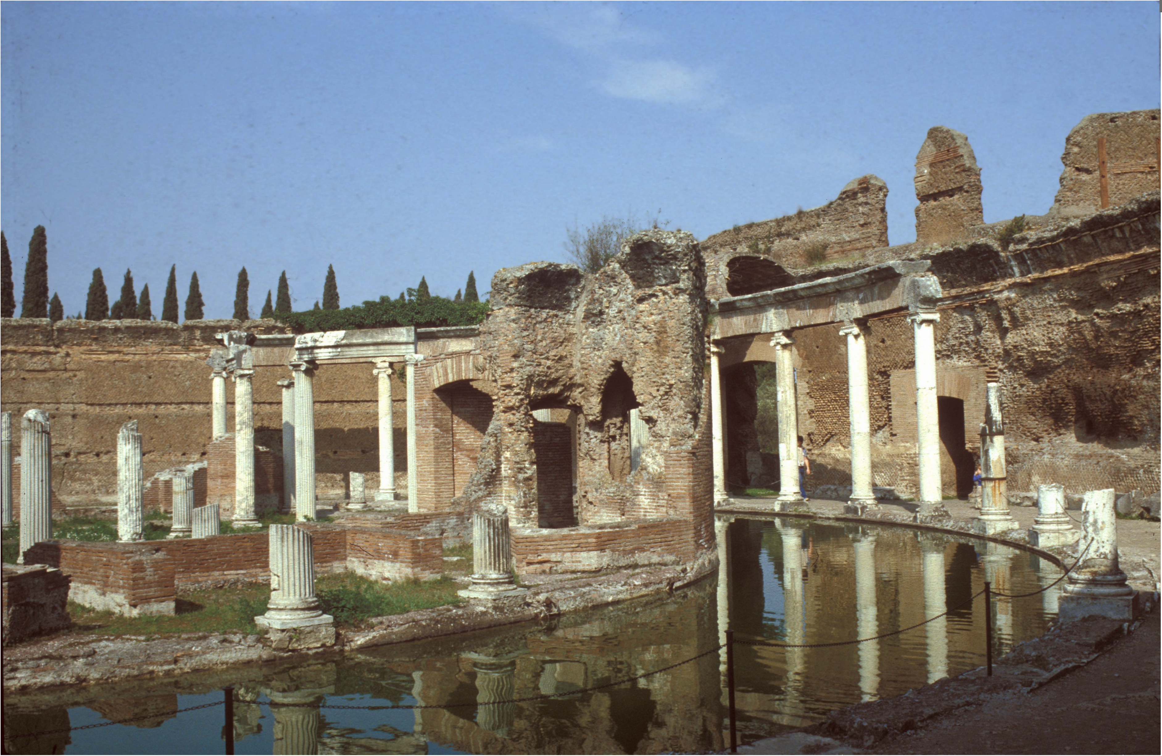 Ruins featuring marble columns, brick walls, and a circular reflecting pool under a blue sky; tall cypress trees line the background.
