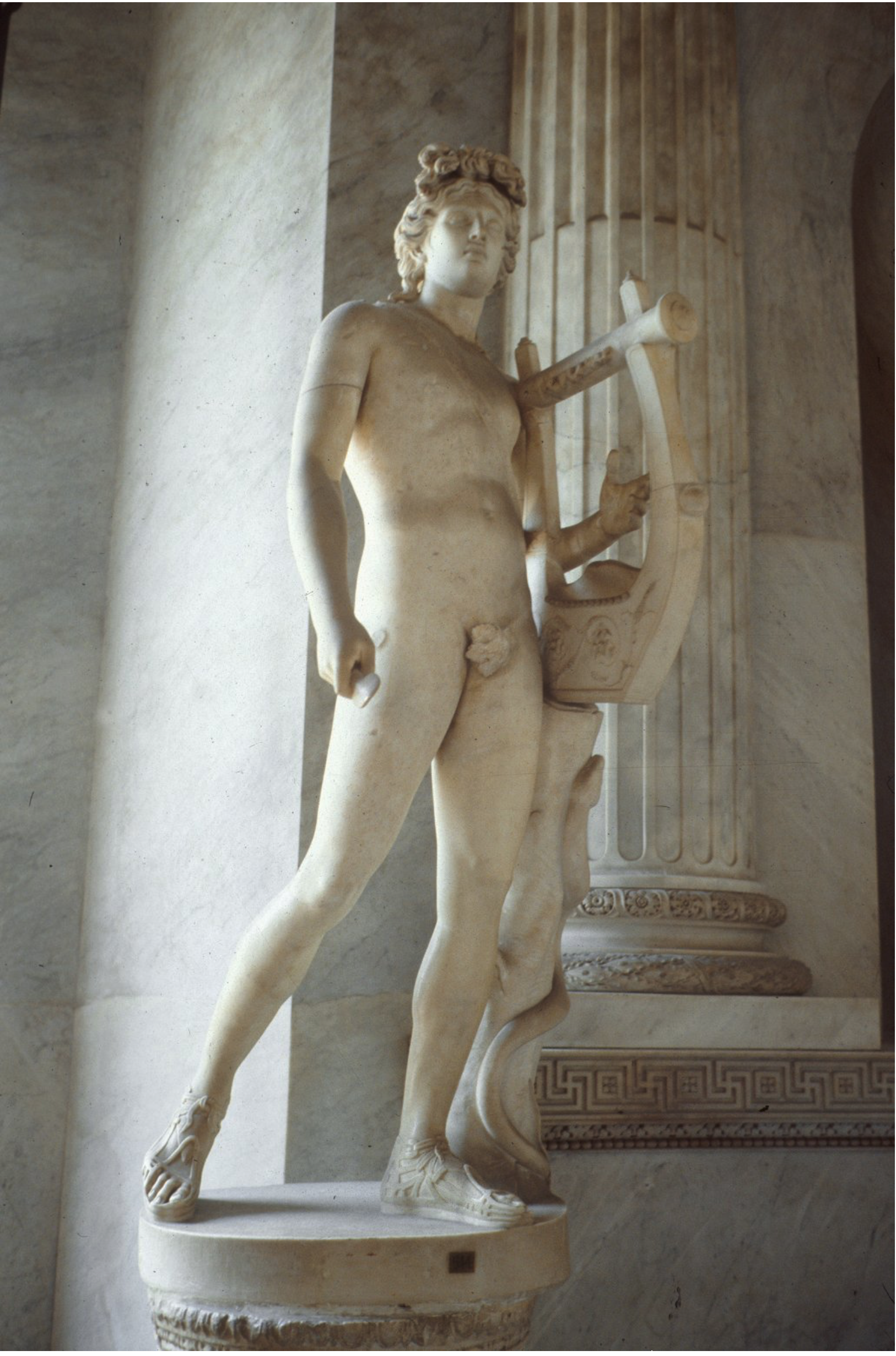 Marble statue of a standing nude male figure with curly hair, holding a lyre. The figure is supported by a decorative branch with a snake and stands on top of a column.