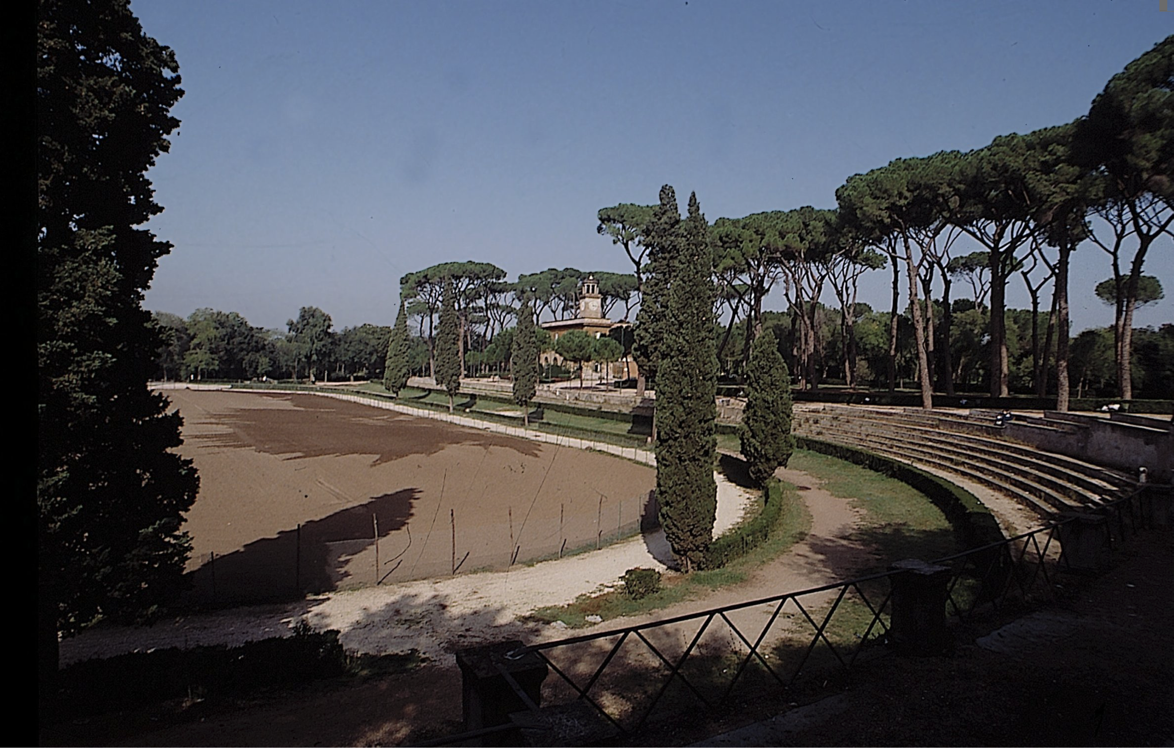 Roman-style stadium with a dirt racetrack arena, surrounded by stone steps and tall cypress and pine trees under a clear blue sky. A small building and greenery are visible in the background.