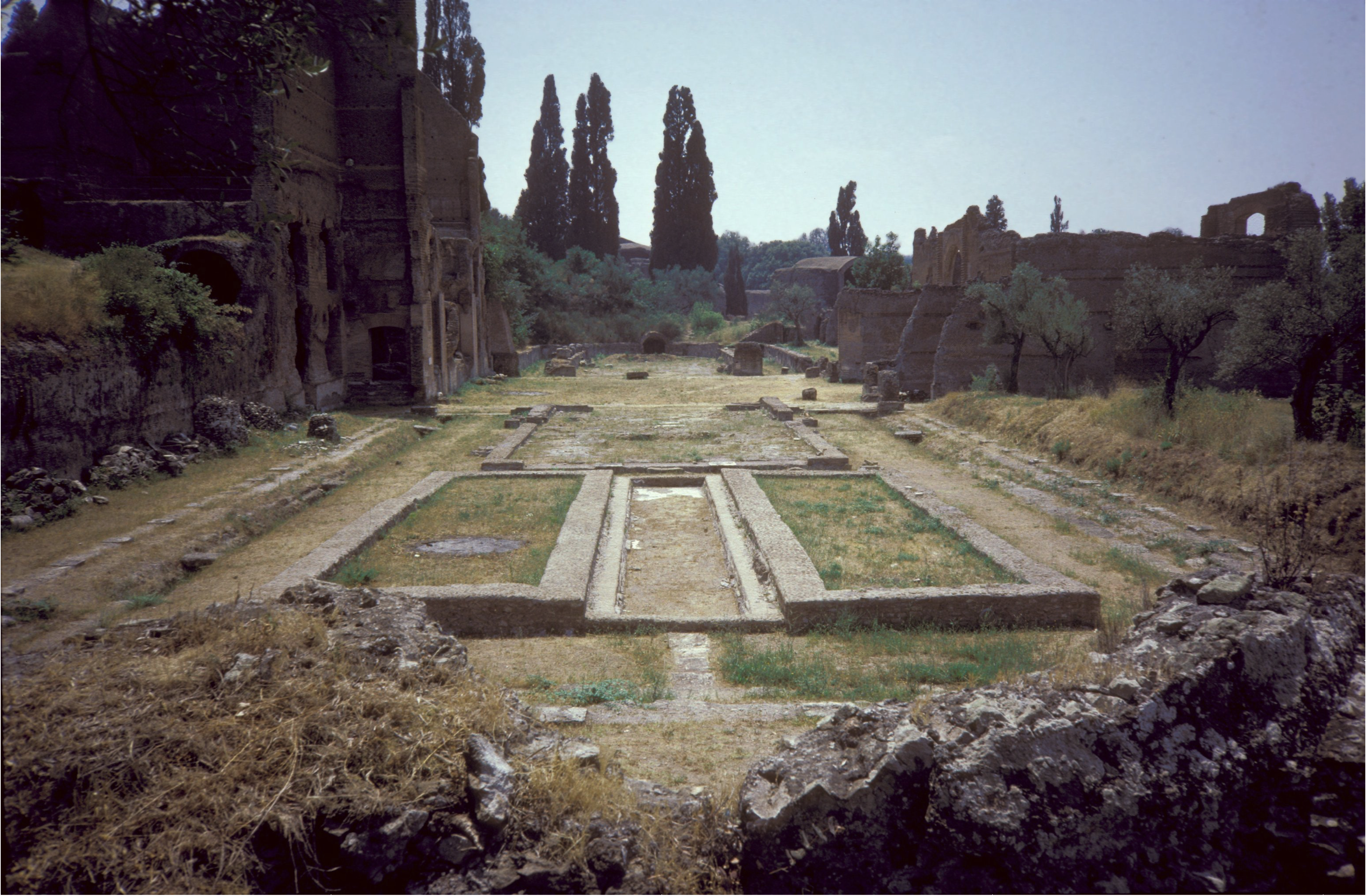 Ruins with long rectangular stone foundations at the center, surrounded by grassy and rocky areas, trees, and the remains of brick structures and walls. Tall cypress trees are visible in the background.