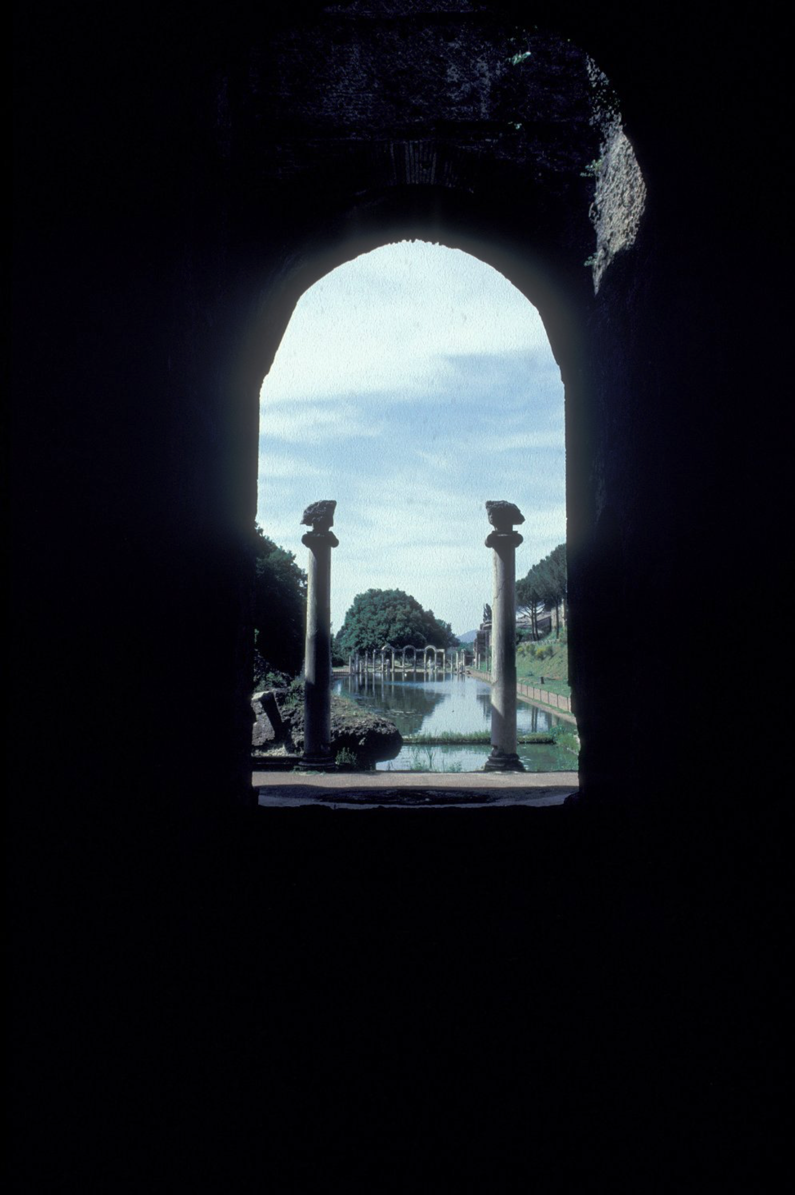 View through a dark arched doorway framing free-standing columns, a reflecting pool, trees, and a pathway.