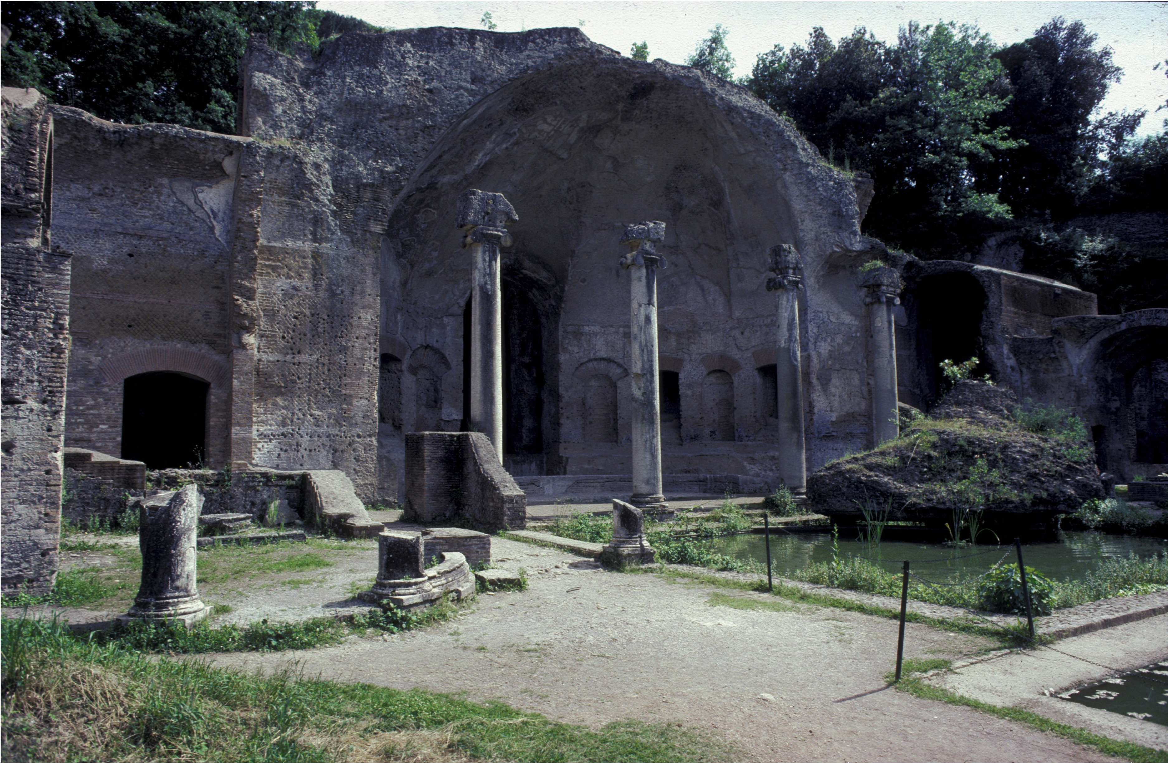 Ruins of a domed brick structure with fragmented walls, and free-standing columns. Overgrown ground vegetation is interspersed with pathways and reflecting pools.
