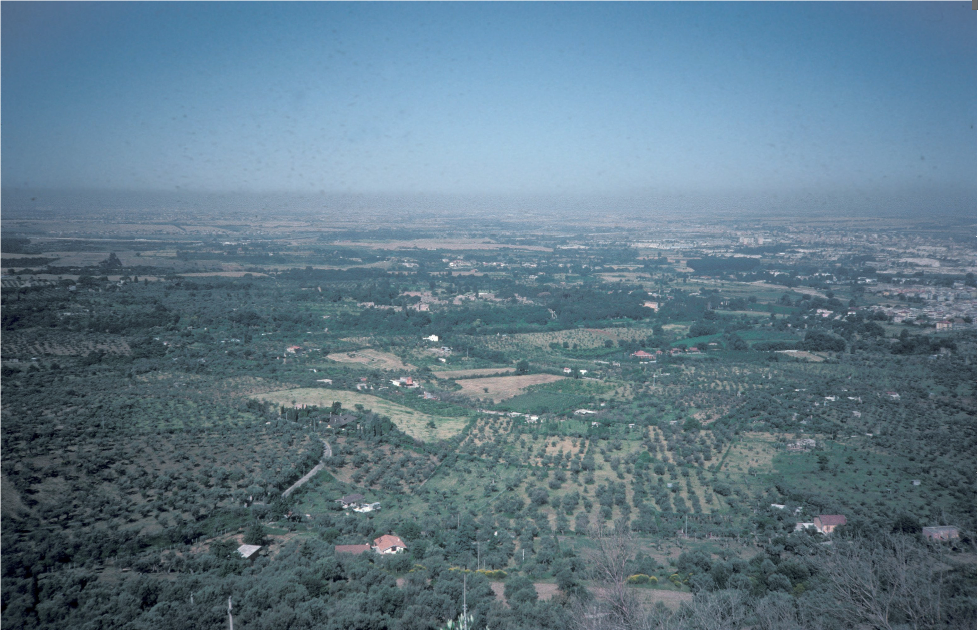 A wide view of a rural landscape with green fields, scattered trees, small buildings, and farmland under a clear blue sky, stretching towards the horizon.