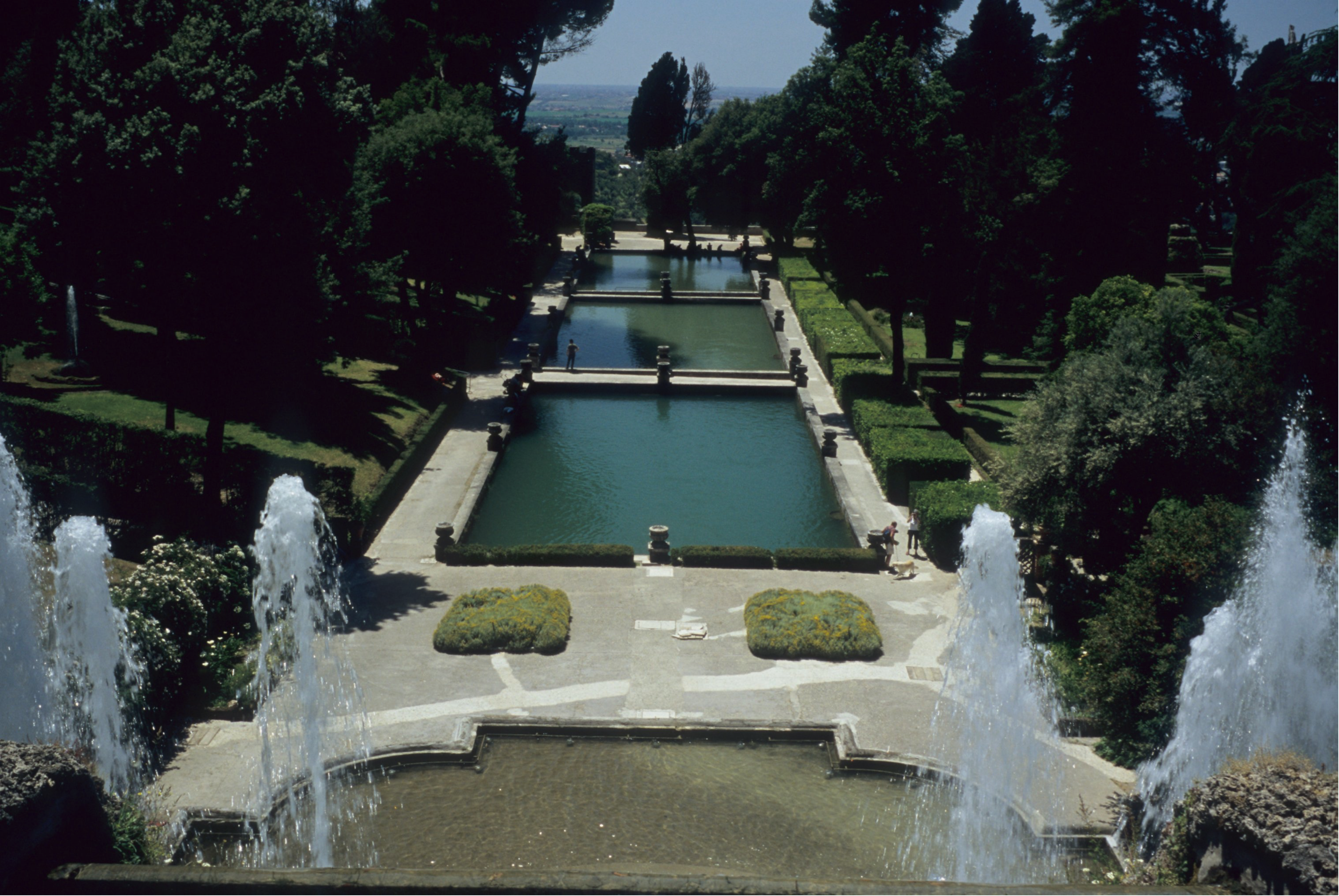 A view from above of a series of rectangular reflecting pool is surrounded by low hedges, decorative urns, and stone pathways. In the foreground, a series of fountains spray water into a shallow fountain pool below.