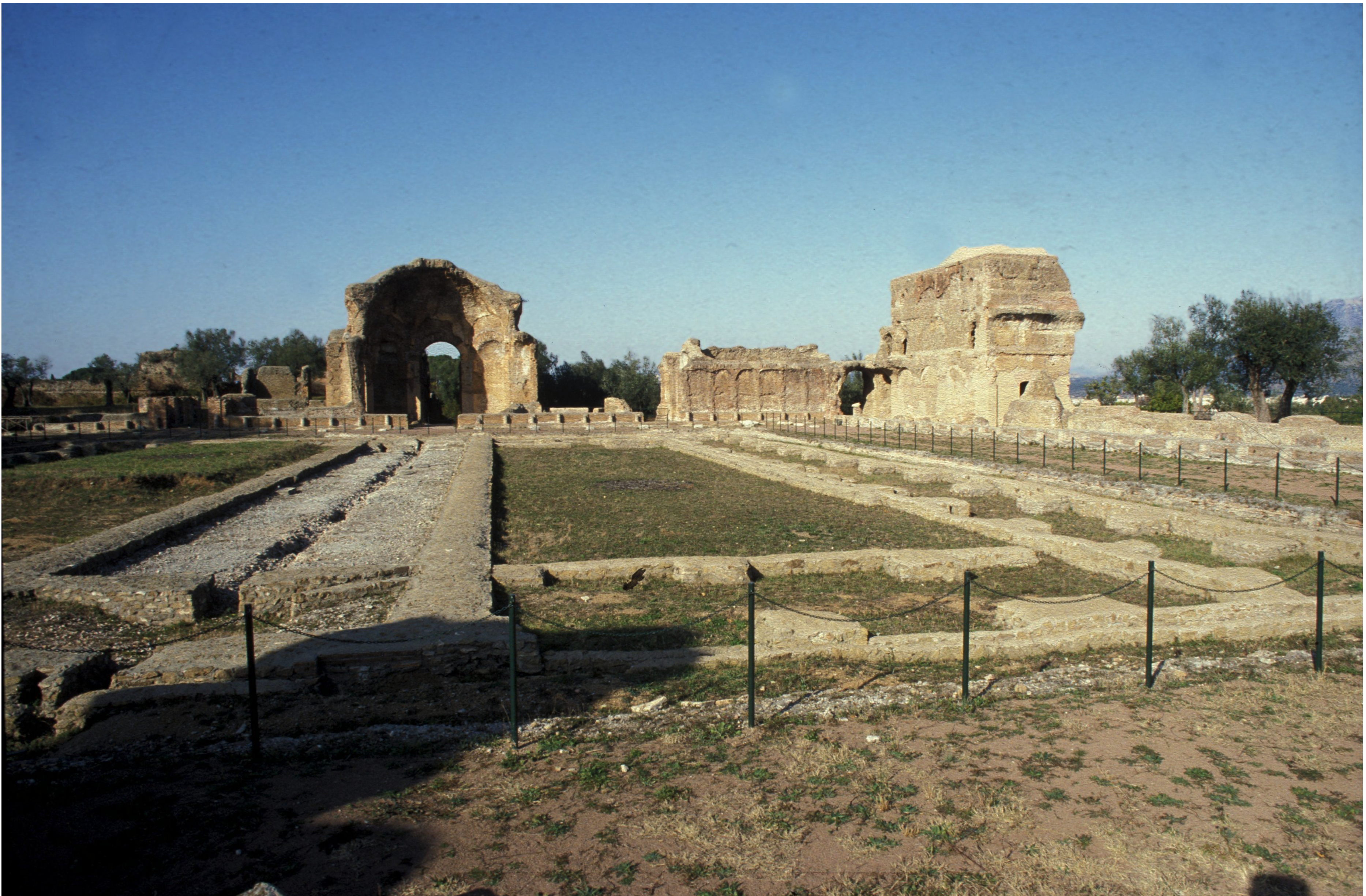 Stone ruins with domed structure, partial walls, and stone foundations surrounded by grass and pathways, enclosed by a low fence. Trees and distant hills are visible under a clear blue sky.