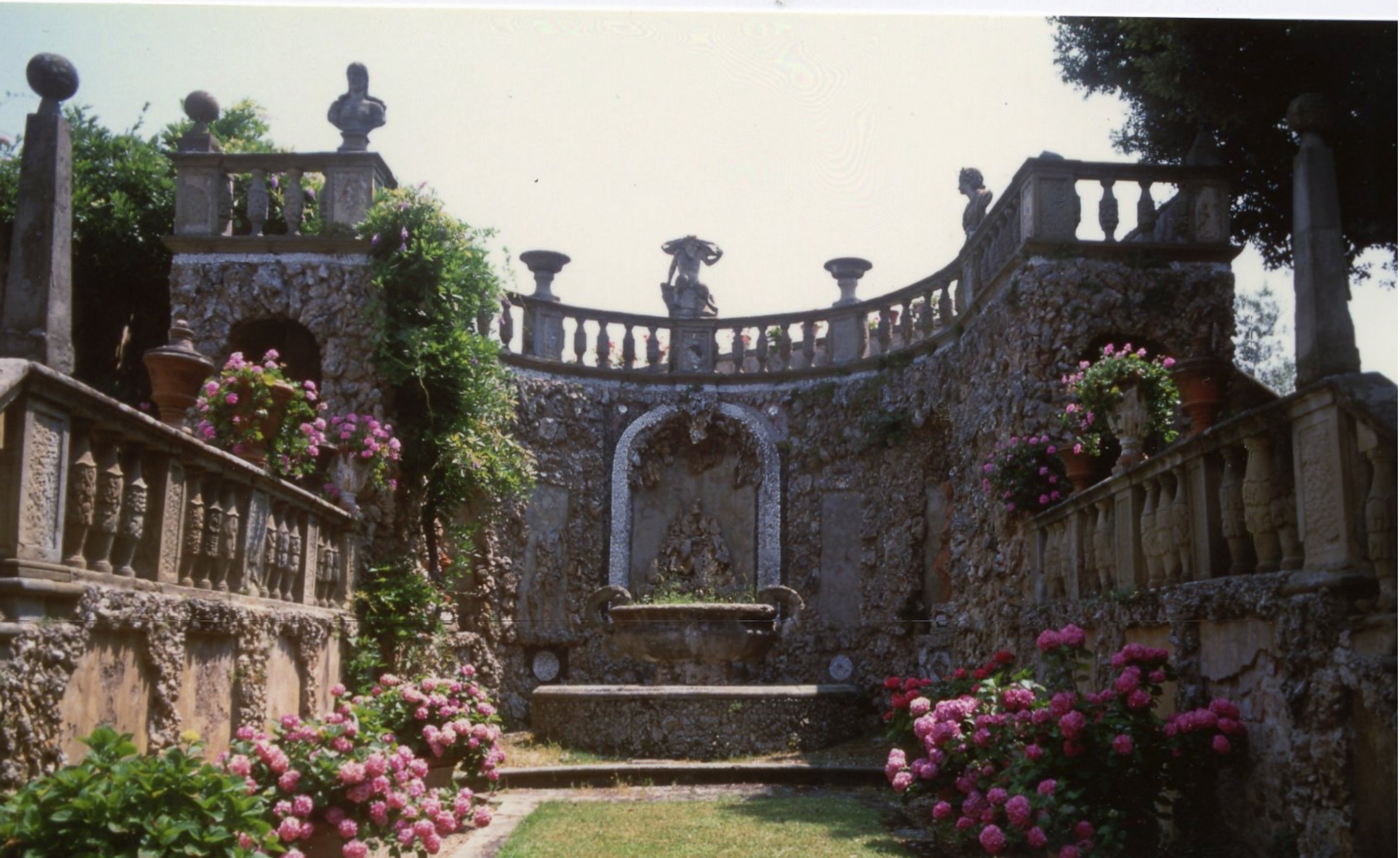 A formal garden with stone walls, terraces, balustrades, and urns filled with pink flowers.