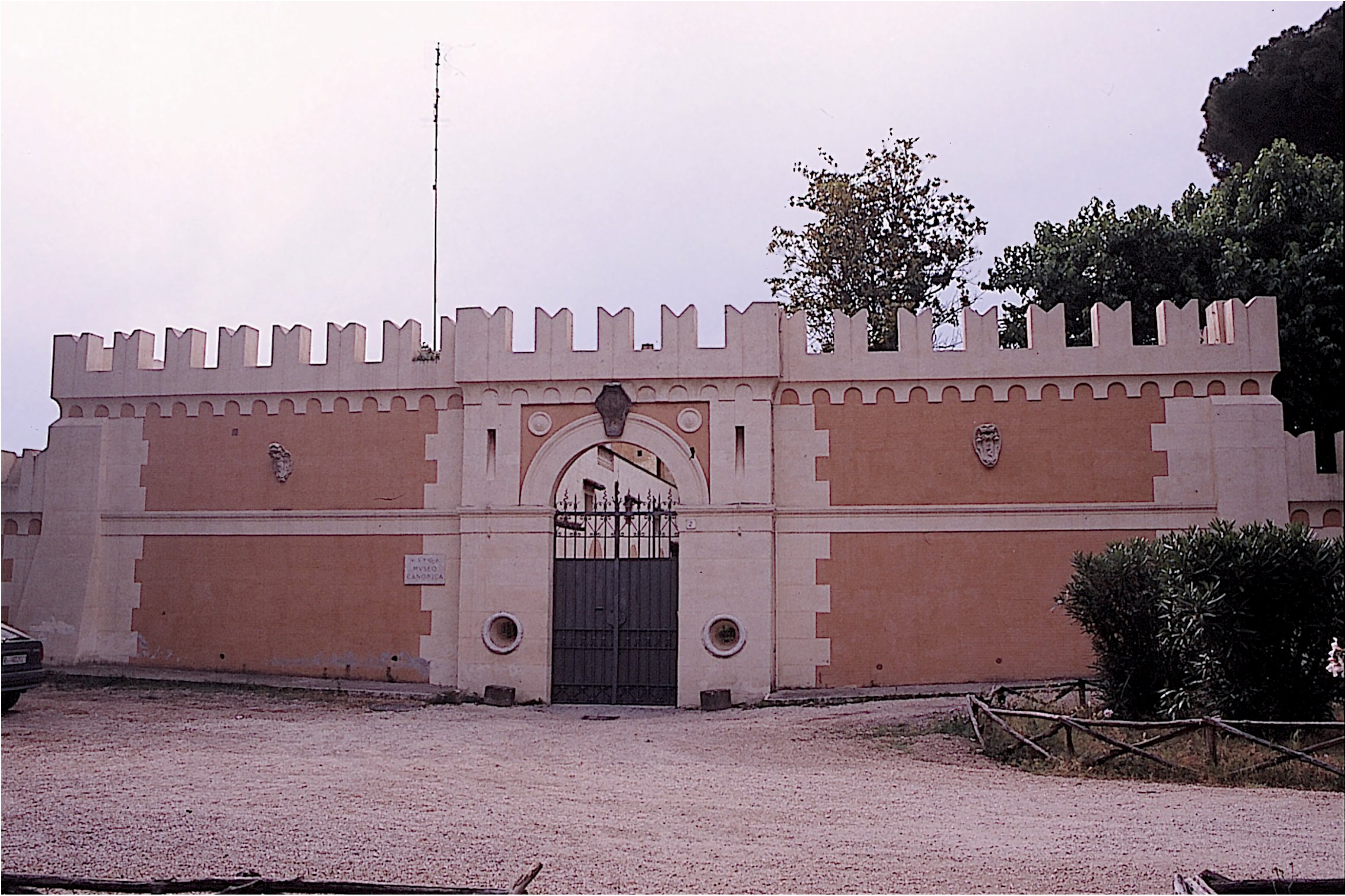 A stone and brick fortress-like structure with crenellated walls, arch, and metal gale, surrounded by trees and bushes. There is a carved stone sign near the entrance and a gravel path in front.