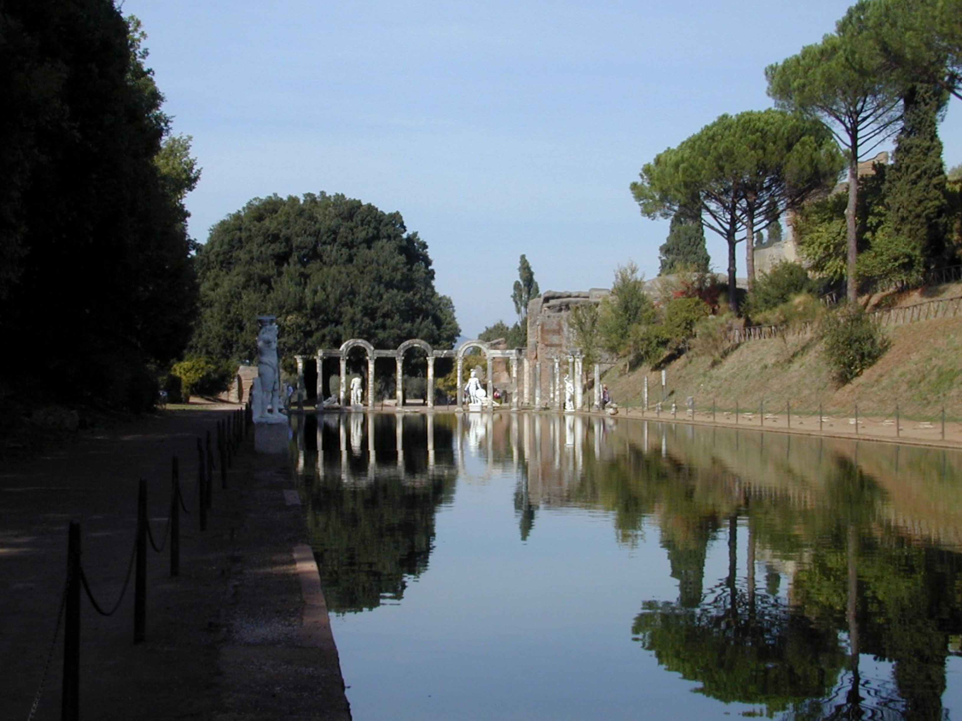 A long rectangular pool is bordered by statues and free-standing arches and columns ruins, with tall trees and greenery reflected in the water.