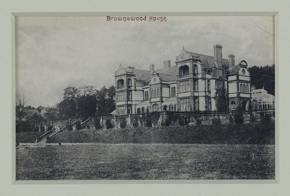 Grand, 4-story building with many large windows. There is a glass conservatory visible on the right side and the photograph is taken from a large lawn in front of the building.
