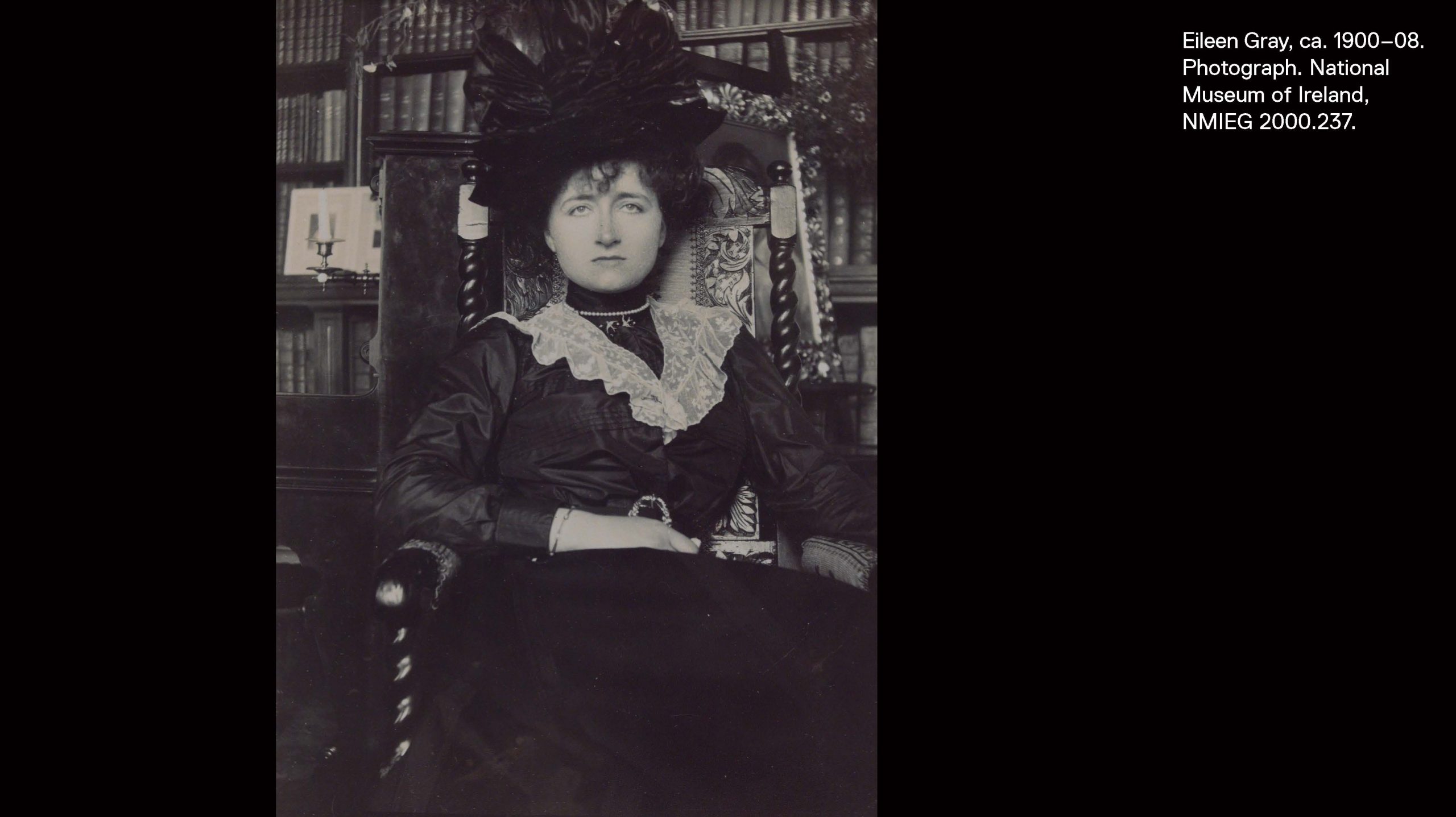 Eileen Gray in Edwardian dress and a dark hat seated in a high-backed chair staring directly at the camera.