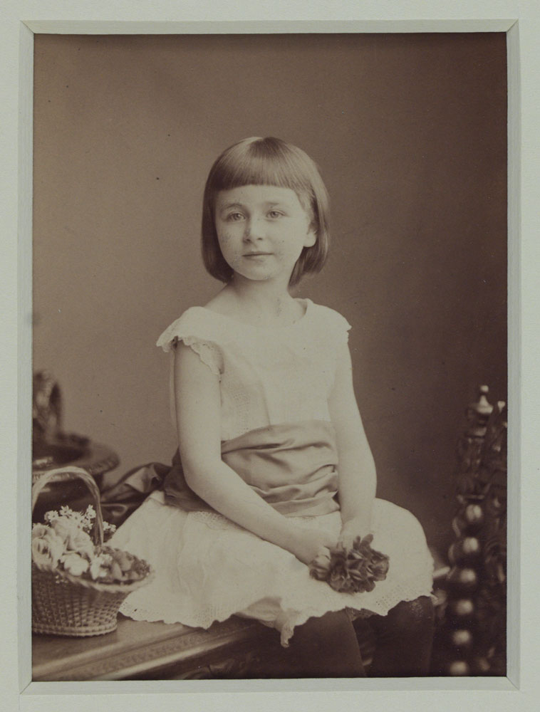 Young girl with chin-length hair wearing a short-sleeved lace dress with a ribbon at the waist sitting on a table. She is holding a flower in her hand and there is a basked of flowers to her right.