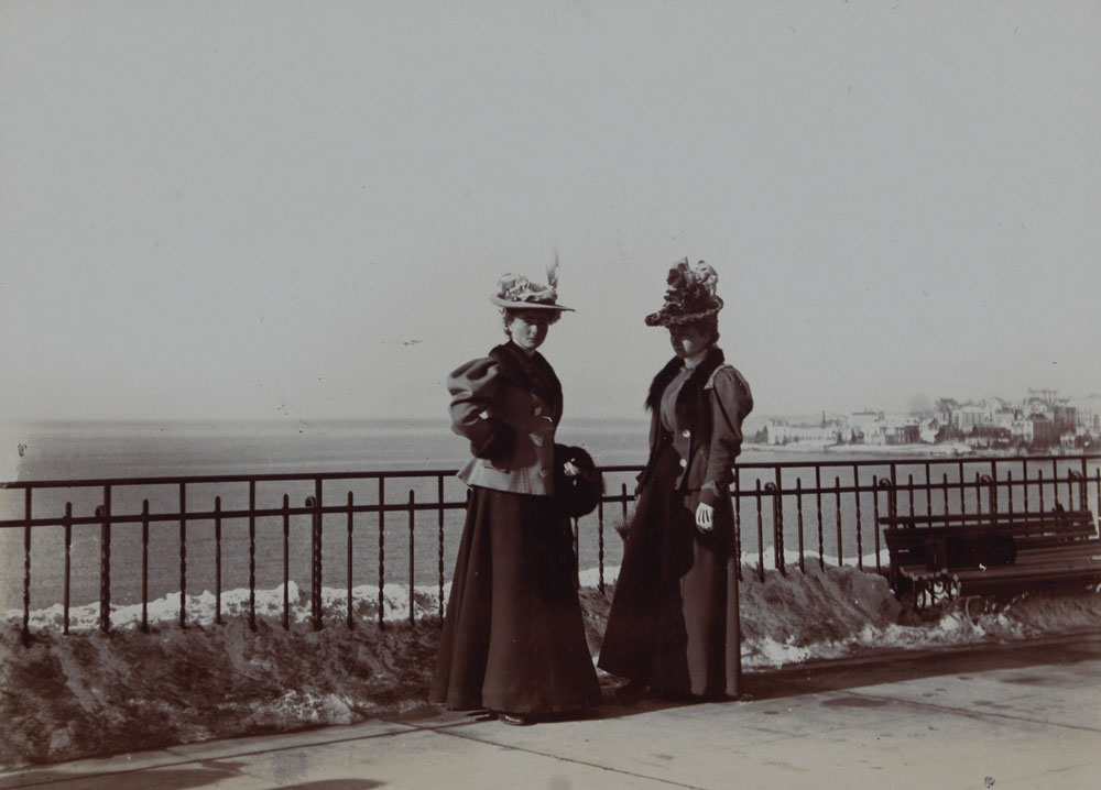 Two young women in 19th-century dress wearing long skirts and fall hats standing next to an iron fence overlooking a body of water with a city in the background.