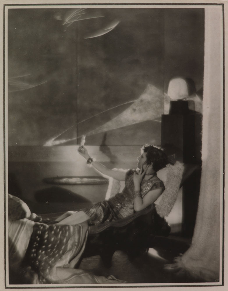 Photograph of woman lounging on a chaise admiring herself in a hand mirror.
