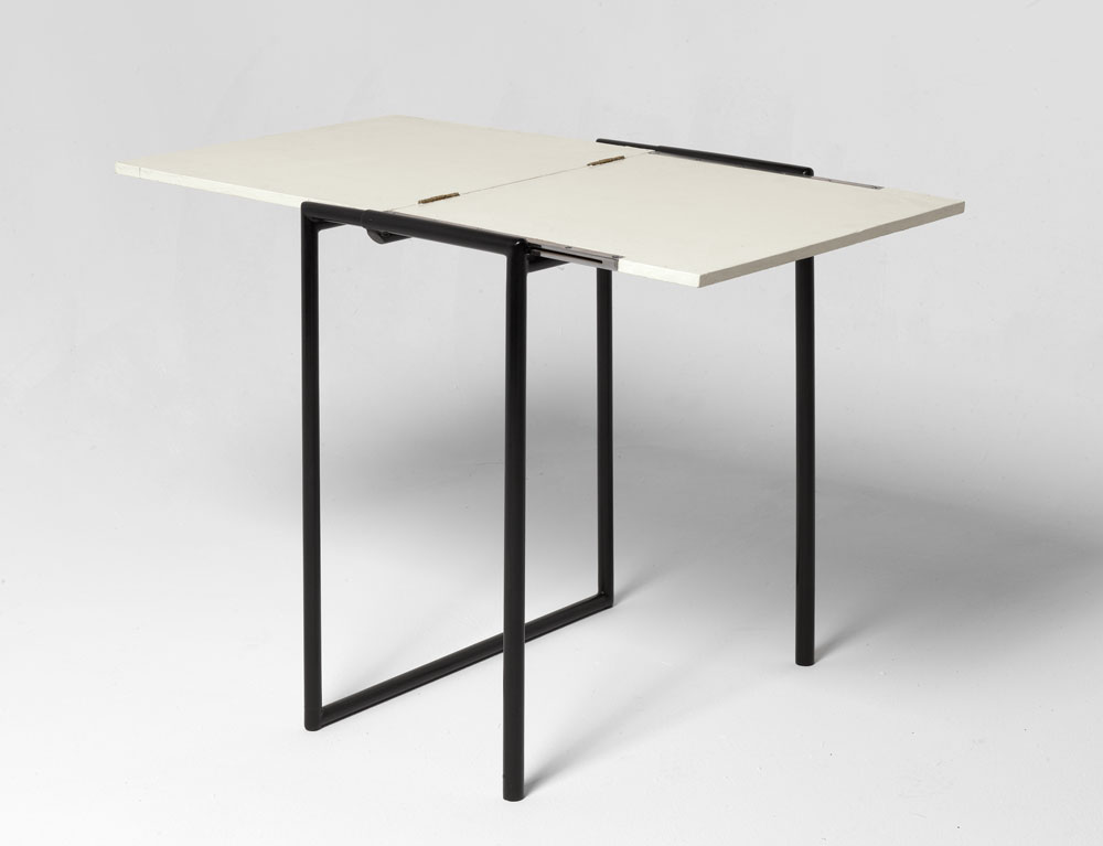 Angular table with black legs and a white top. Two of the black tube legs are connected at the floor and the table top has a center hinge that enables the user to fold it up or extend it.