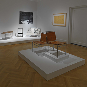 Exhibition image of two Eileen Gray chairs in the foreground and three other pieces of furniture in the background.