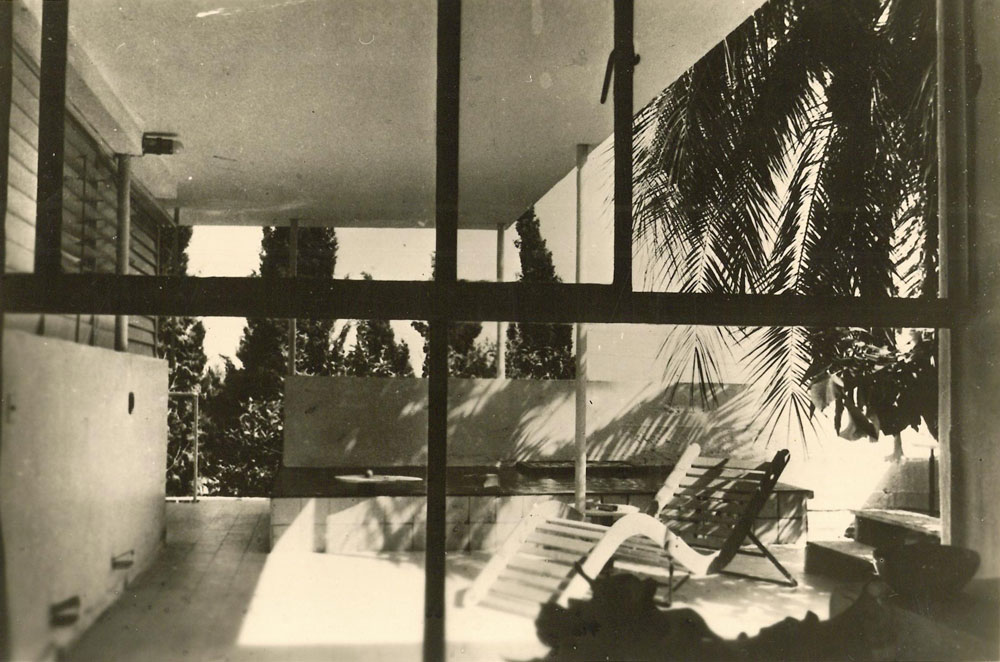 Interior view through a wall of windows to a covered terrace with deck chairs and trees.