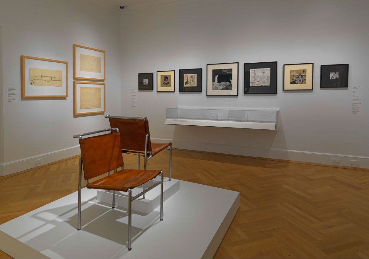 Exhibition image of two chairs and multiple works on the wall.