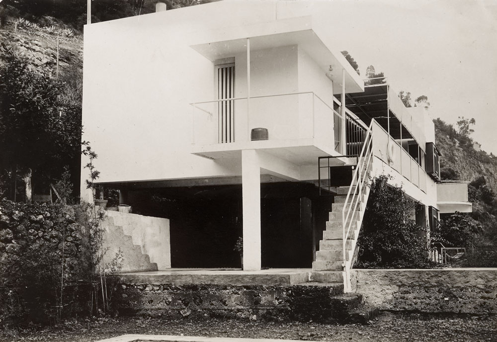 Exterior view of a white modern home raised off the ground with stilts.
